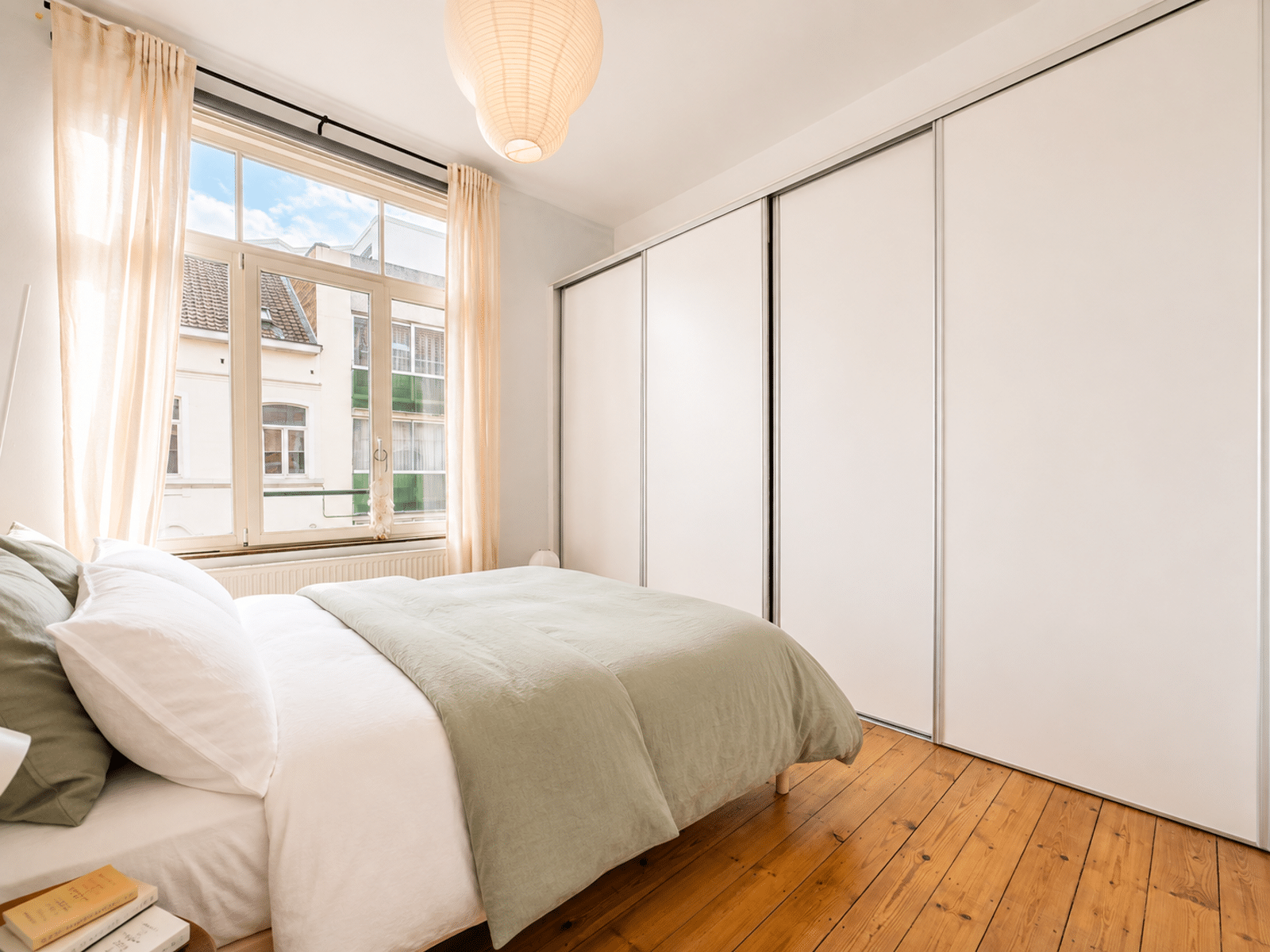 Chambre Lumineuse avec Rangements – You Real Estate Agency Chambre lumineuse d’appartement avec parquet, lit blanc, placards intégrés et grande fenêtre.