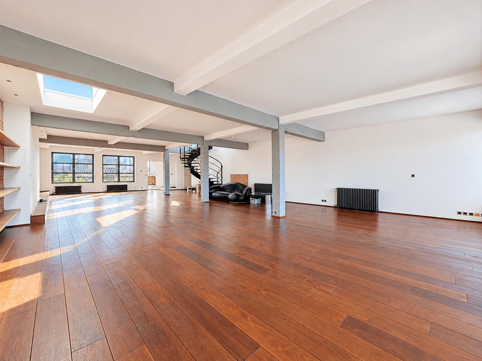 whise photo 16 Spacious loft with wooden floors, gray beams, and large windows letting in sunlight; spiral staircase and seating area in the distance.