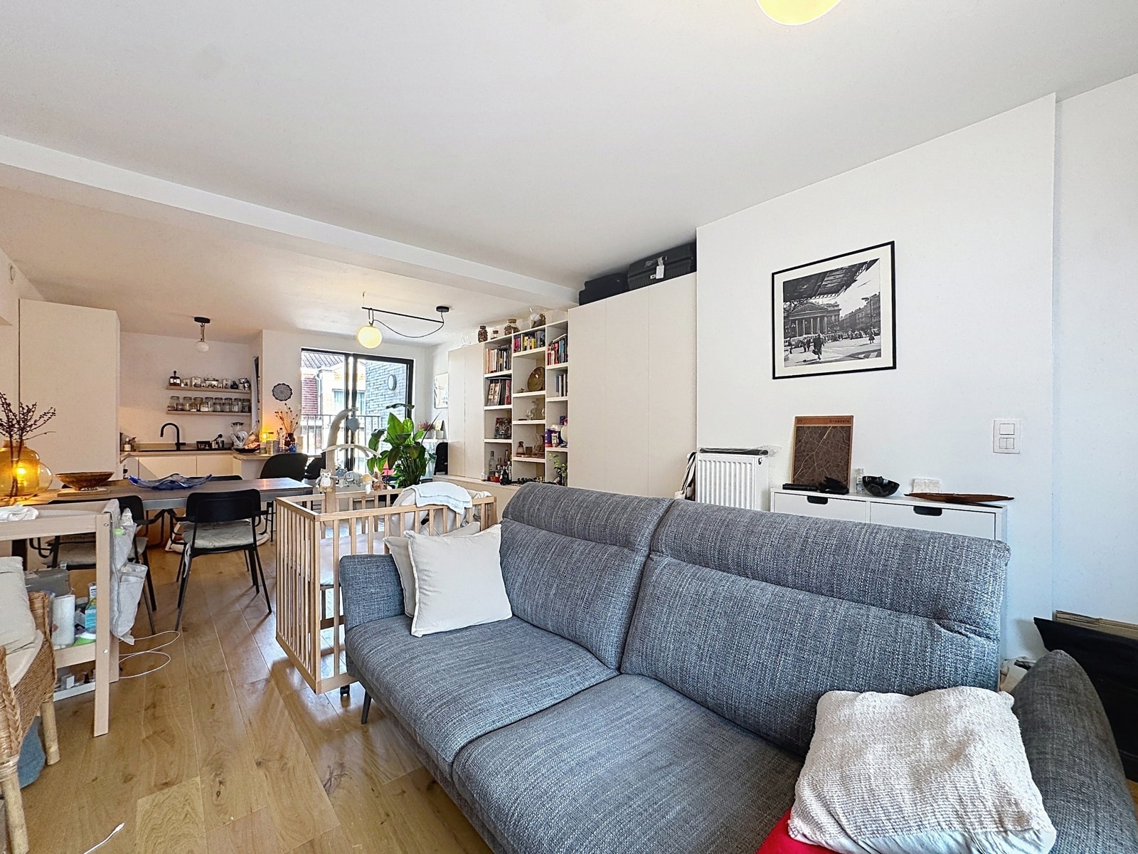 whise photo 5 Open-plan living area with a gray sofa in the foreground, wooden floor, and a dining table toward the back, plus a kitchen and shelves. Plants and warm lighting create a cozy home interior as seen from the seating area.