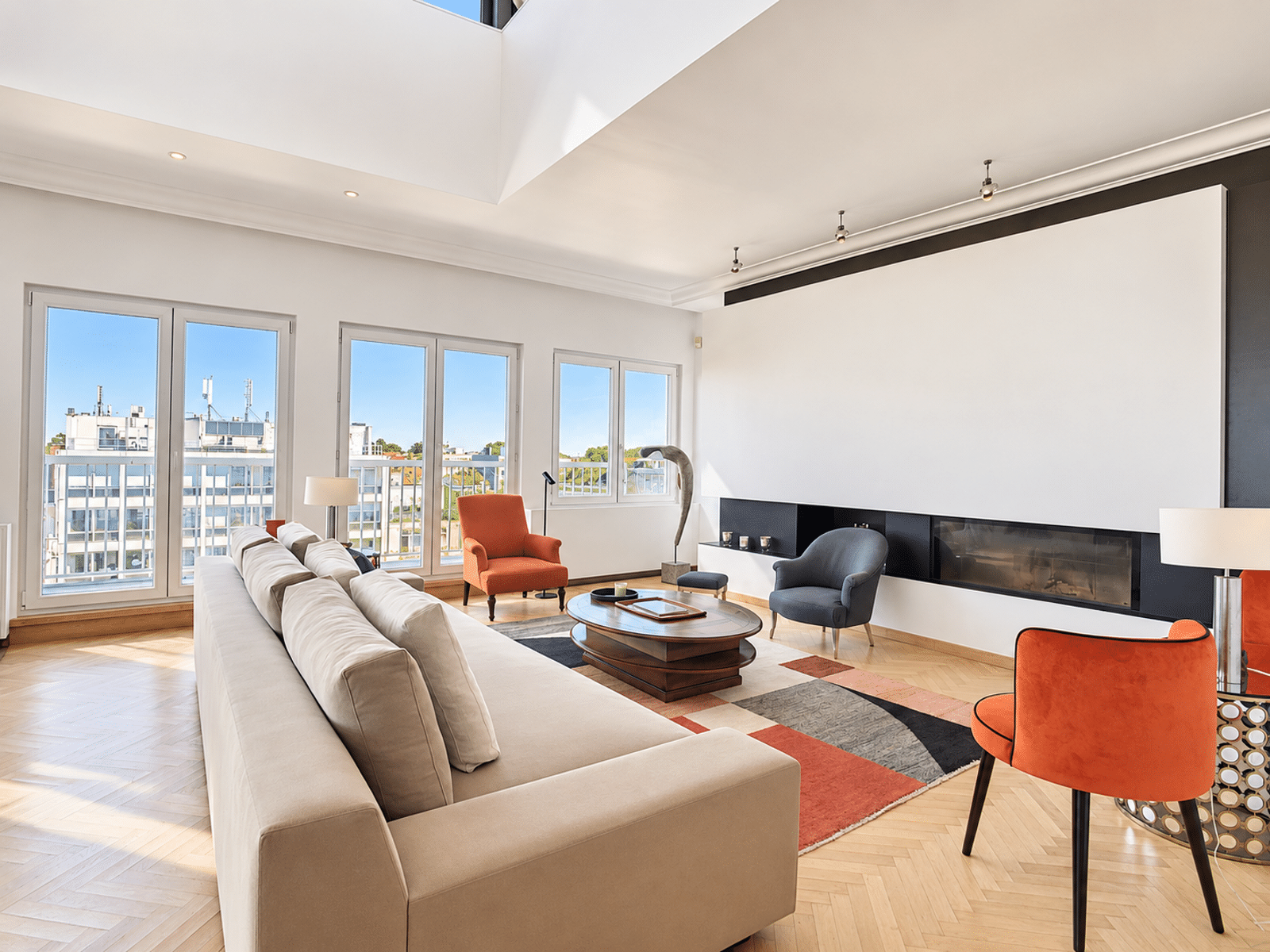 Salon Moderne avec Cheminée – You Real Estate Agency Bright modern living room with a beige sectional, orange chairs, a round wood coffee table, and large windows overlooking a cityscape.