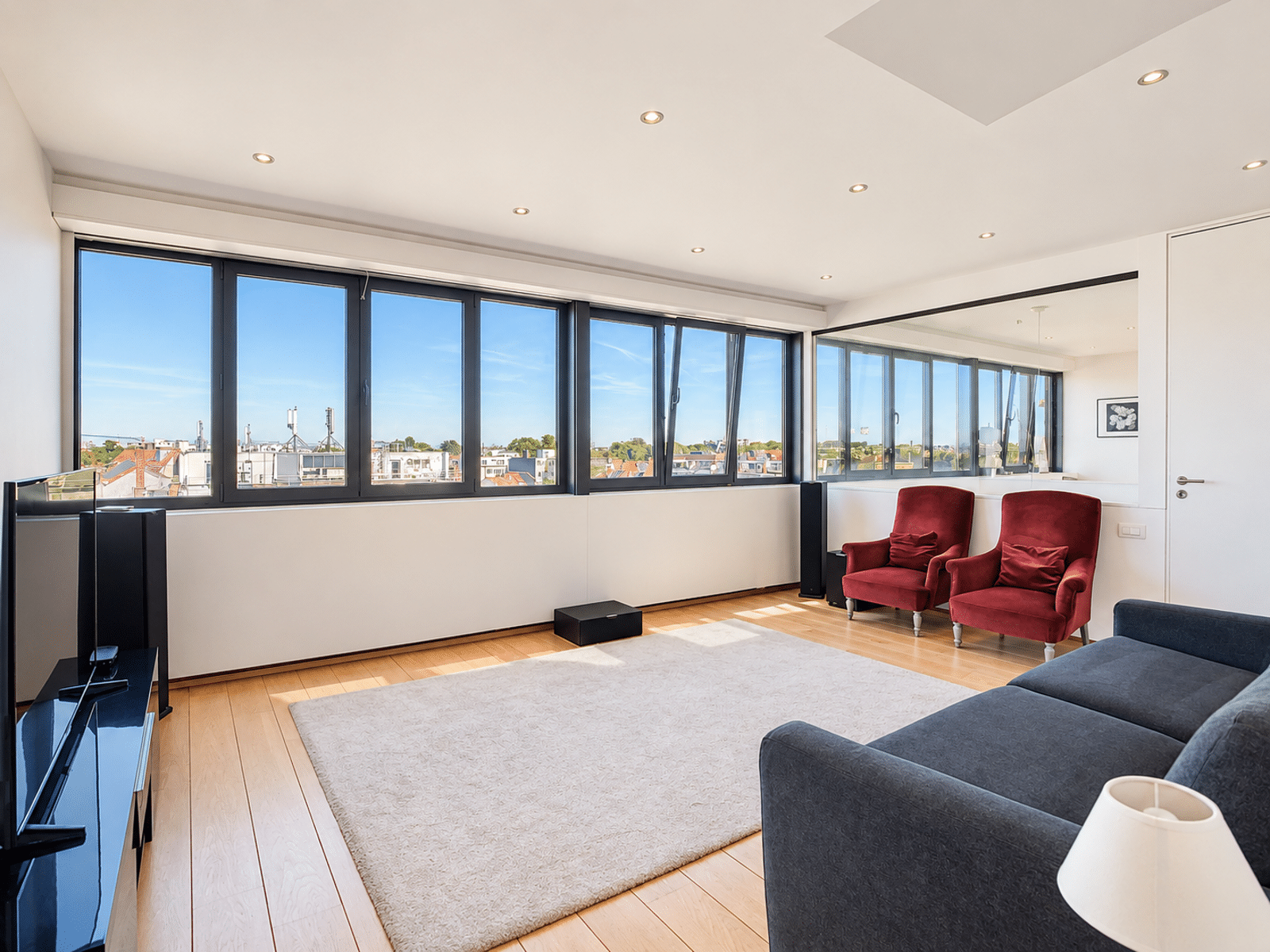 Salon Moderne avec Grandes Fenêtres – You Real Estate Agency Bright modern living room with large panoramic windows and a light beige rug, red chairs, a dark sofa, and wooden floors.