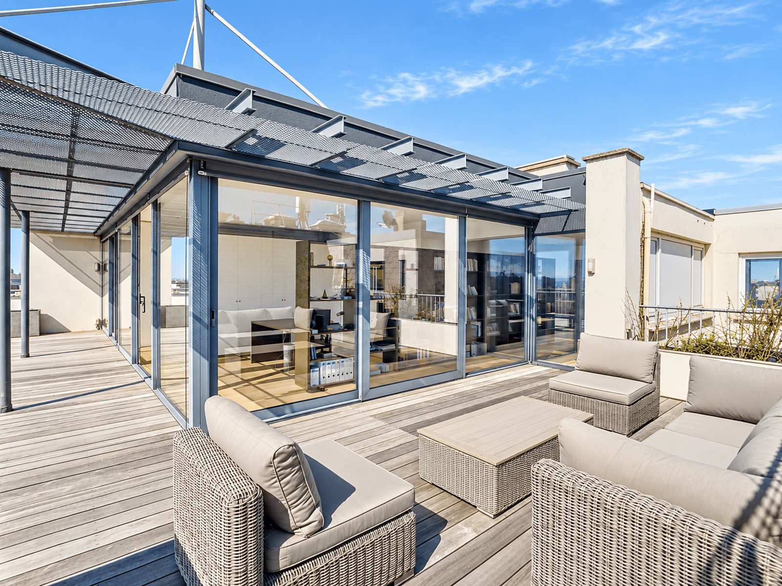 whise photo 2 Rooftop terrace with a glass-walled office module and beige wicker seating on a wooden deck under a clear blue sky.