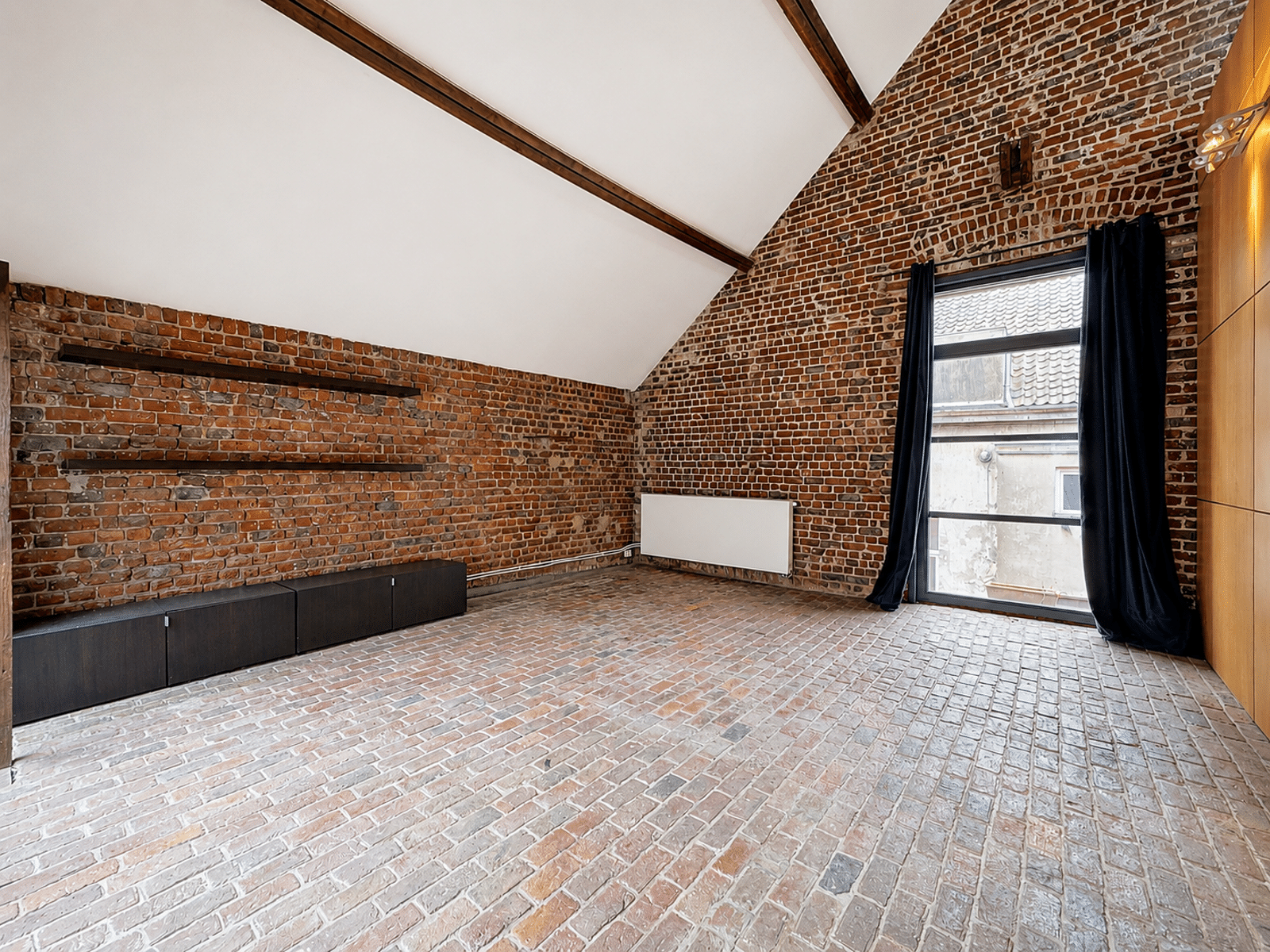 Salon Loft en Briques Apparentes – You Real Estate Agency Brick-walled loft interior with exposed wooden beams, low dark cabinets along the left wall, and a large window with dark curtains.