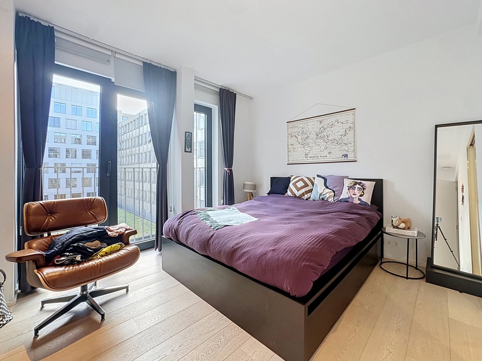 whise photo 11 Modern bedroom with a purple duvet bed against a white wall, tall windows with dark curtains, and a brown leather chair by the window