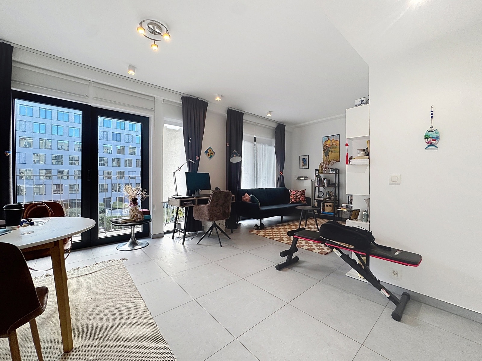 whise photo 6 Bright living space with large glass doors to a city view, a study desk, and a dark sofa set beside a gym bench in the foreground.