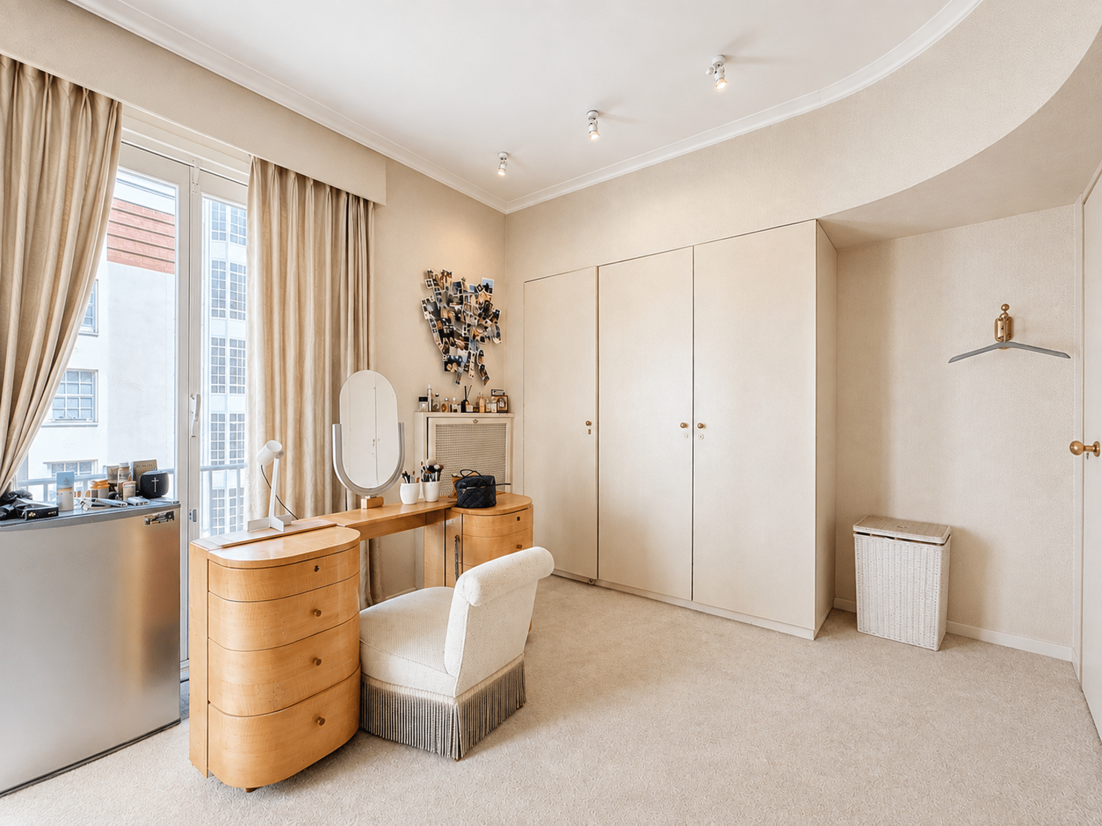 Chambre avec Dressing et Coiffeuse – You Real Estate Agency Vanity desk with rounded drawers and oval mirror, makeup tools on the surface, by a large window with beige curtains