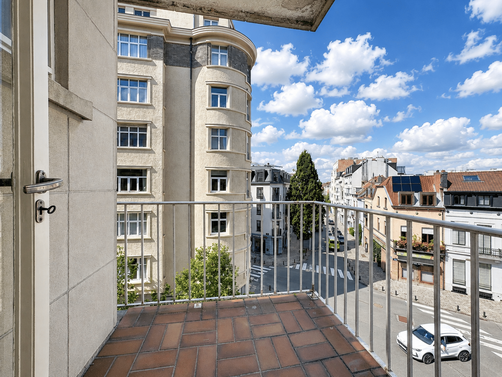 Balcon avec Vue sur Rue Résidentielle – You Real Estate Agency Balcon d’appartement avec vue sur rue résidentielle et immeubles urbains.
