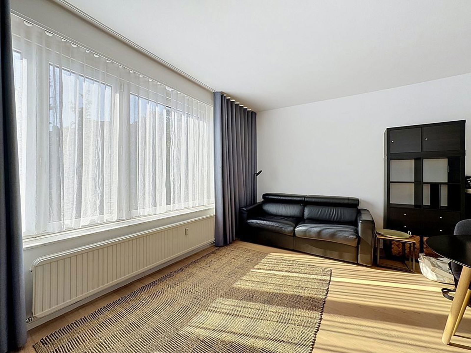Salon Lumineux Avec Grande Baie Vitrée – You Real Estate Agency Salon lumineux d’appartement avec grande baie vitrée, canapé noir et parquet clair.