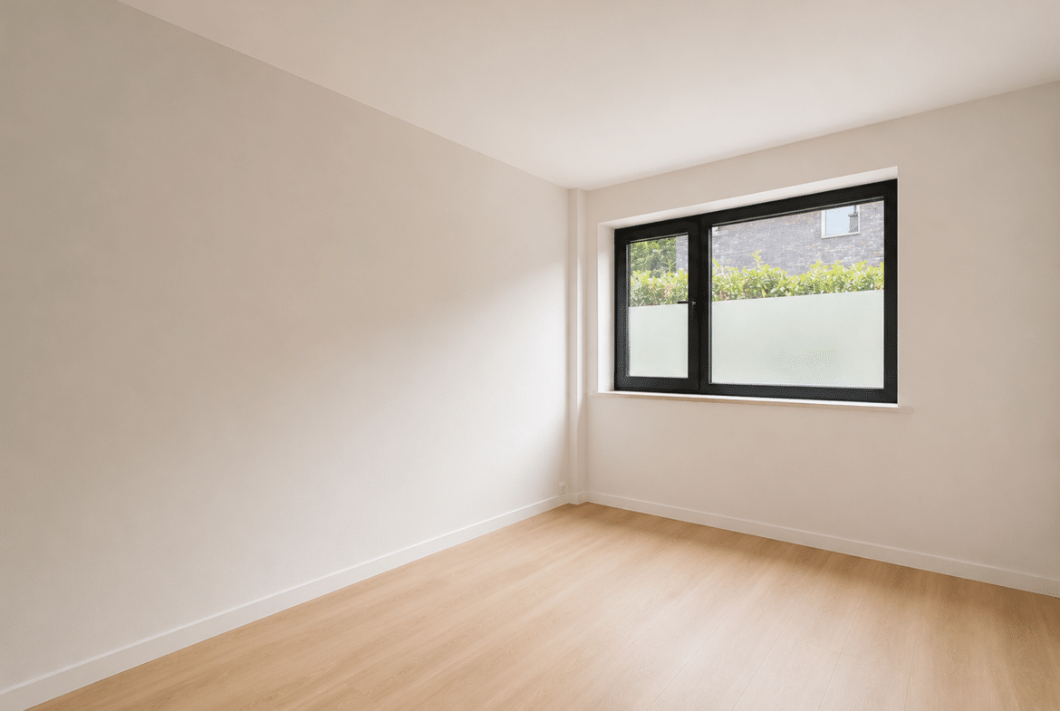 Empty sunlit room with light wood flooring and a large black-framed window showing greenery outside; minimal decor.