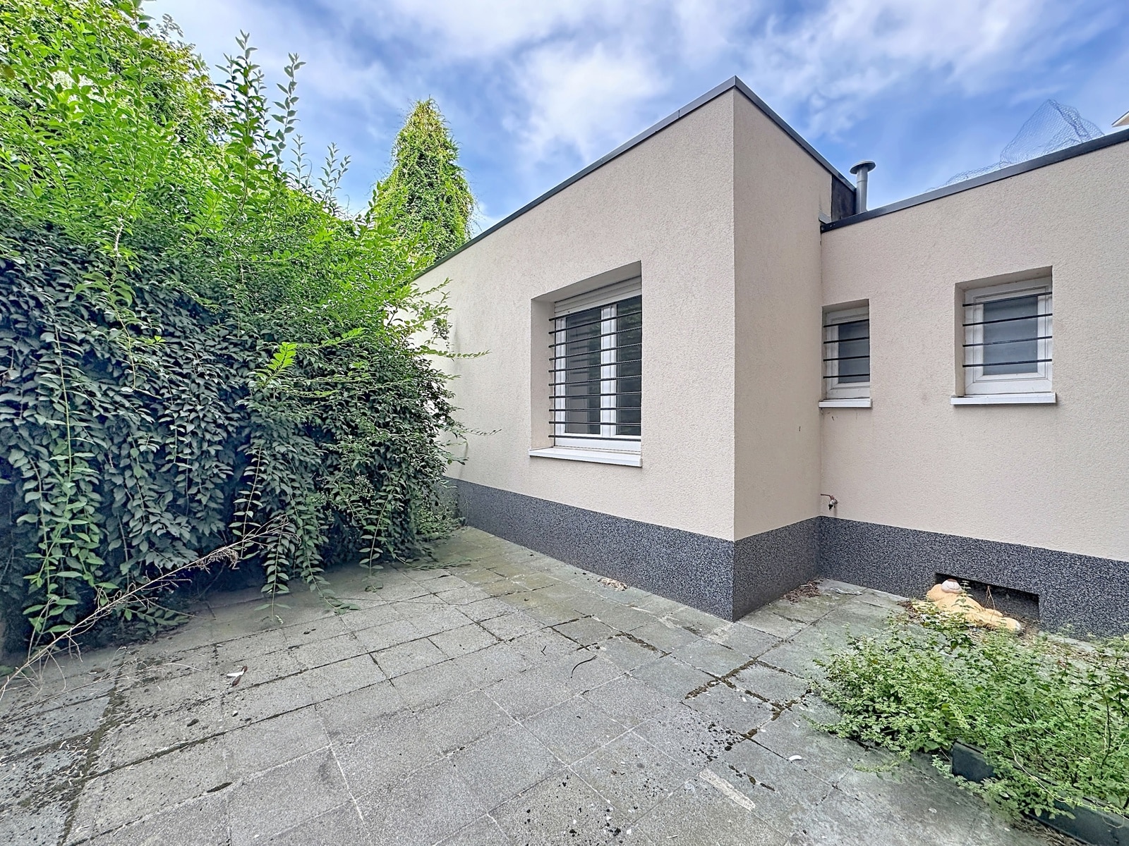 Terrasse Dallée De Maison Moderne – You Real Estate Agency Terrasse extérieure dallée d’une maison moderne avec façade claire et végétation.
