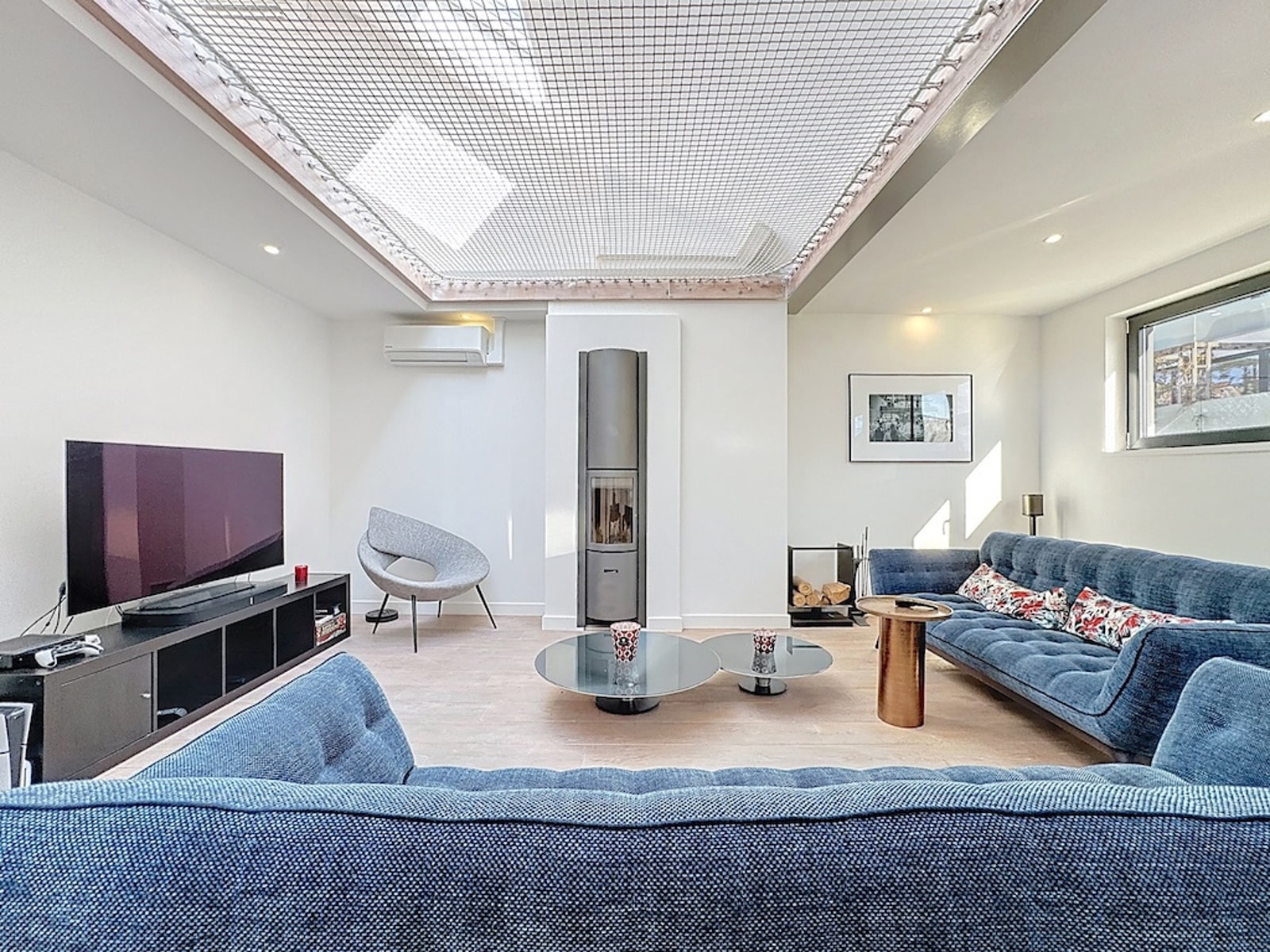 whise photo 14 Bright modern living room with a blue sectional, glass coffee tables, and a wall-mounted TV under a grid skylight ceiling in white walls.