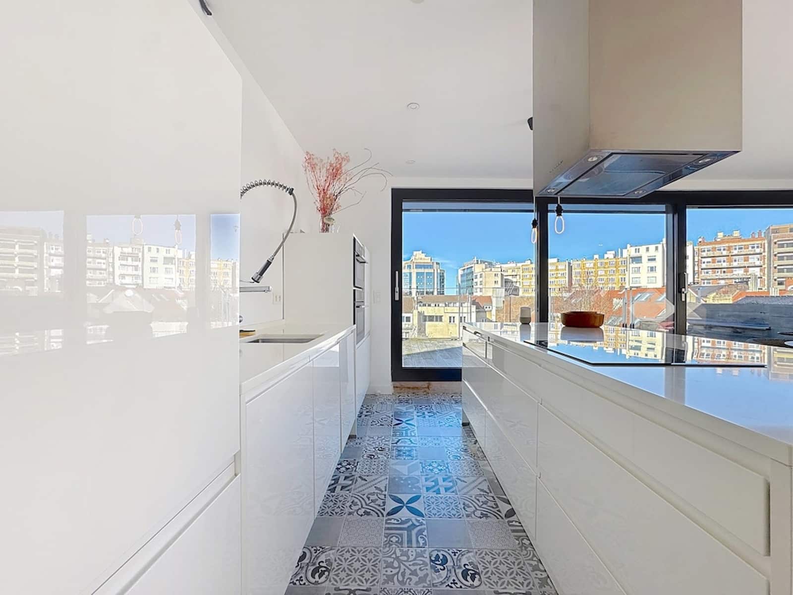 whise photo 7 Bright white kitchen with glossy cabinets, a central island, and large windows showing a cityscape; patterned tiled floor.