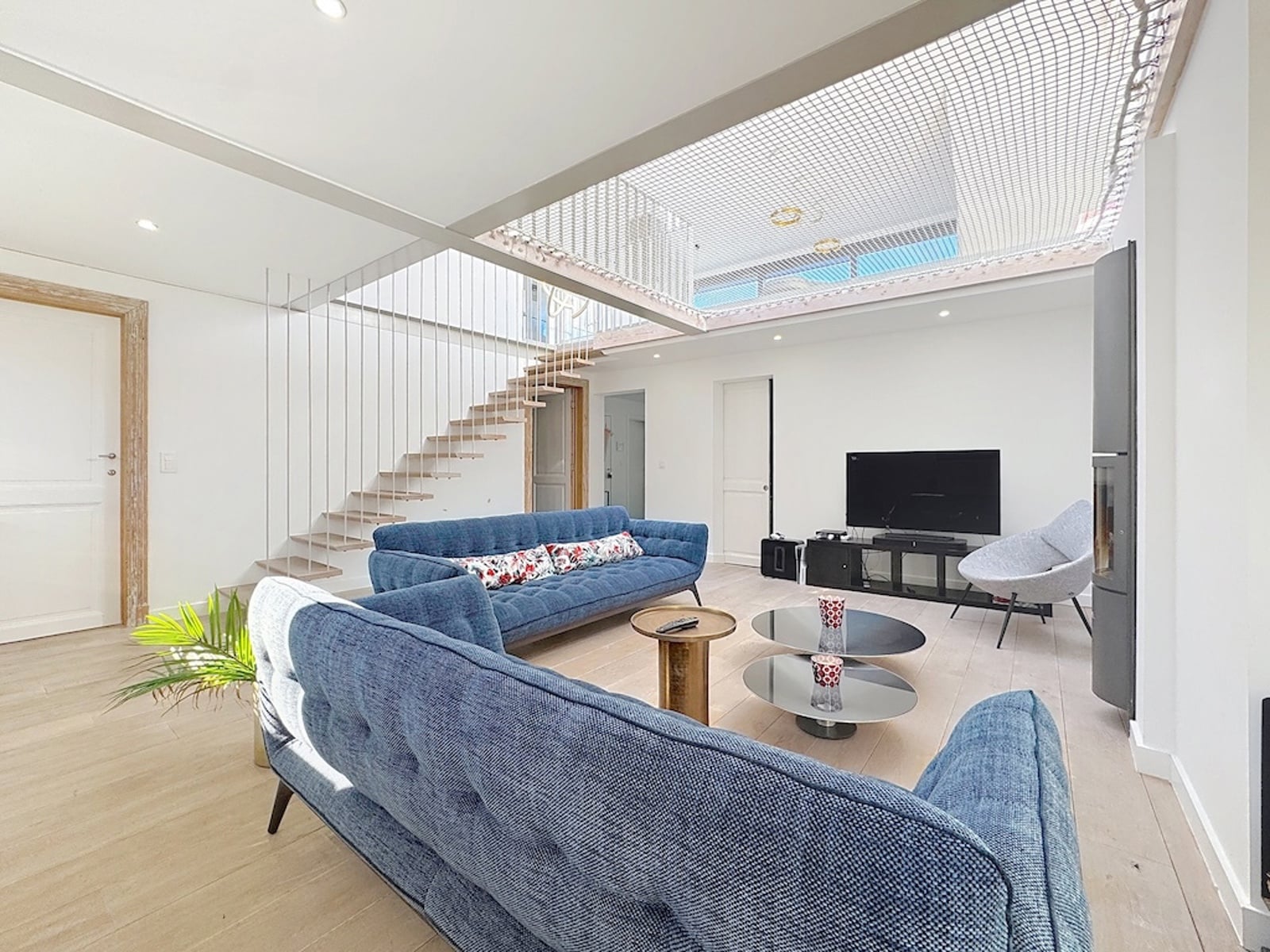 Salon Lumineux avec Mezzanine – You Real Estate Agency Spacious living room with blue upholstered sofas, a coffee table, and a TV setup against a white wall.