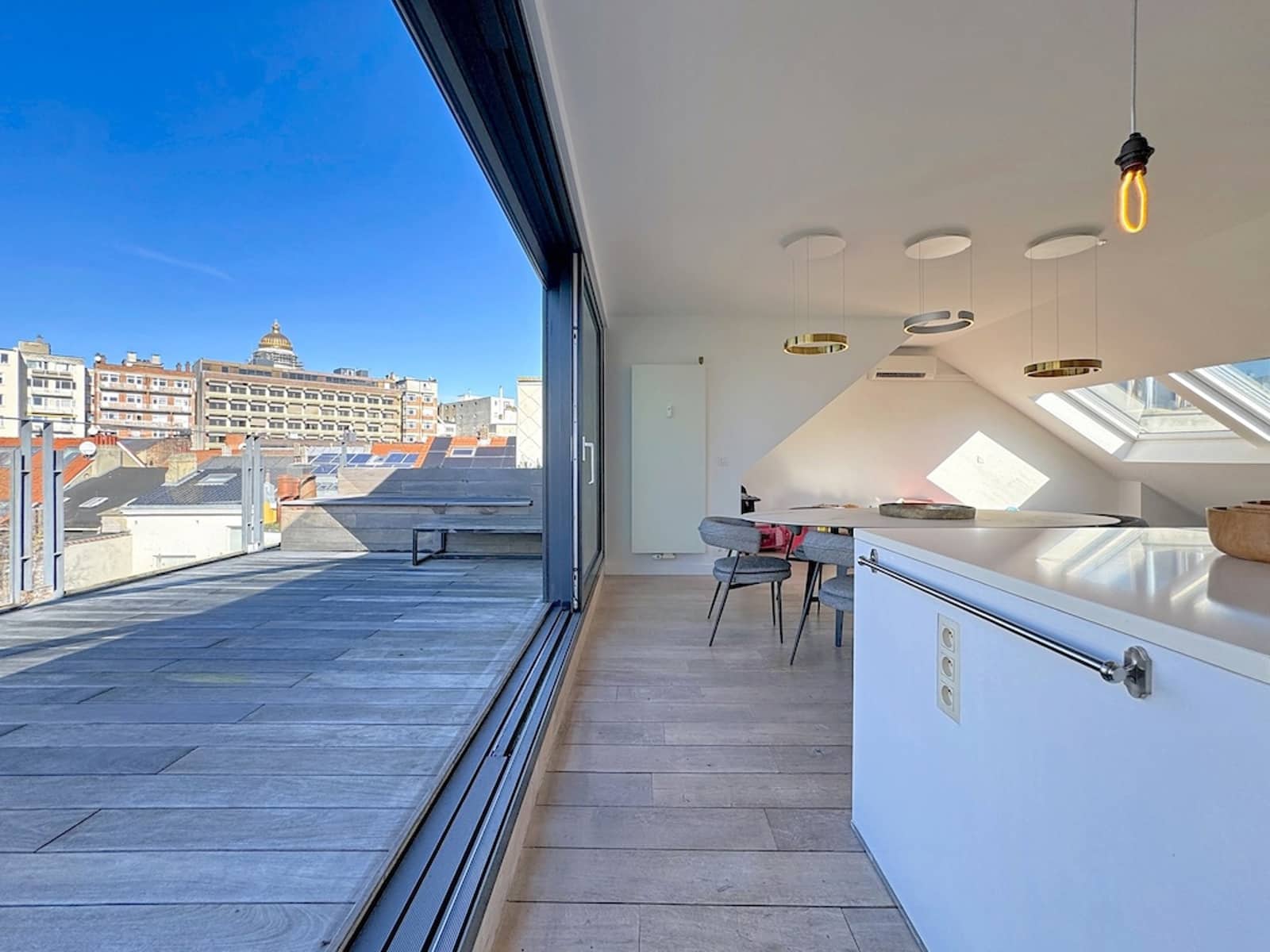 Cuisine Moderne avec Terrasse – You Real Estate Agency Open-plan kitchen/dining with large sliding glass doors to a wooden balcony and a city skyline beyond.