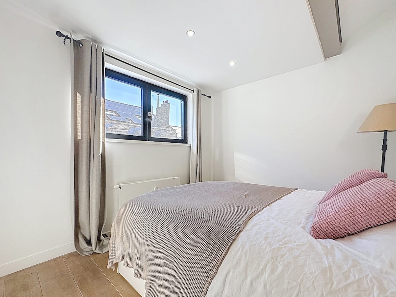 Chambre Lumineuse avec Lit Blanc – You Real Estate Agency Sunlit bedroom with a white bed, beige throw, pink textured cushions, and a window framed by beige curtains.