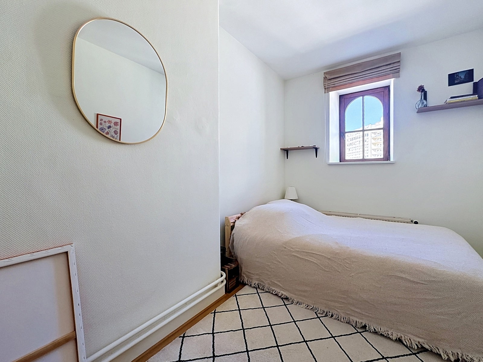 whise photo 15 A small bedroom with a single bed covered in a light blanket, white walls, and a window showing a cityscape outside.