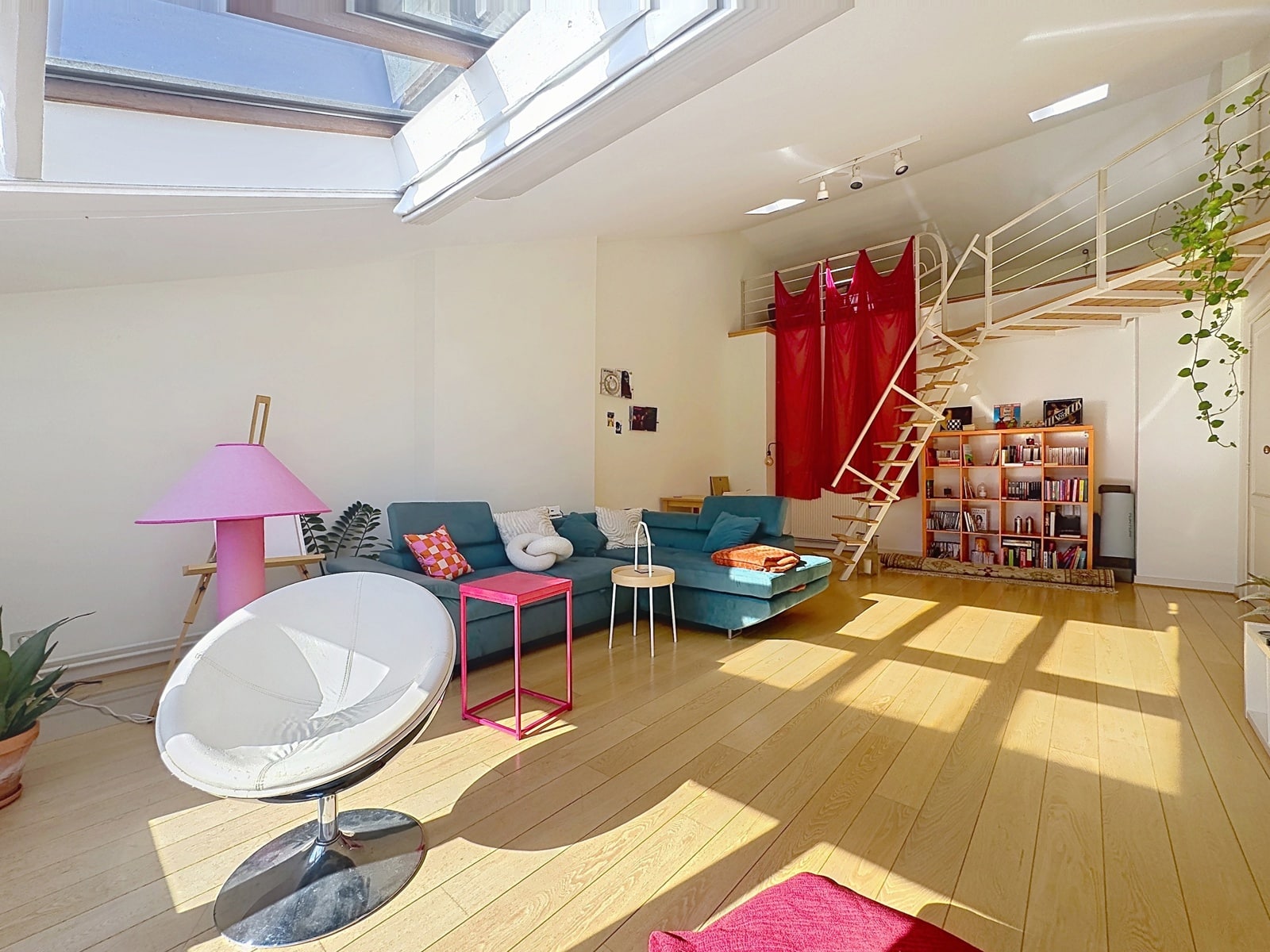 whise photo 3 Bright loft living room with a teal sectional, pink lamp, and round white chair on a light wood floor under skylights; red curtain blocks a mezzanine staircase.