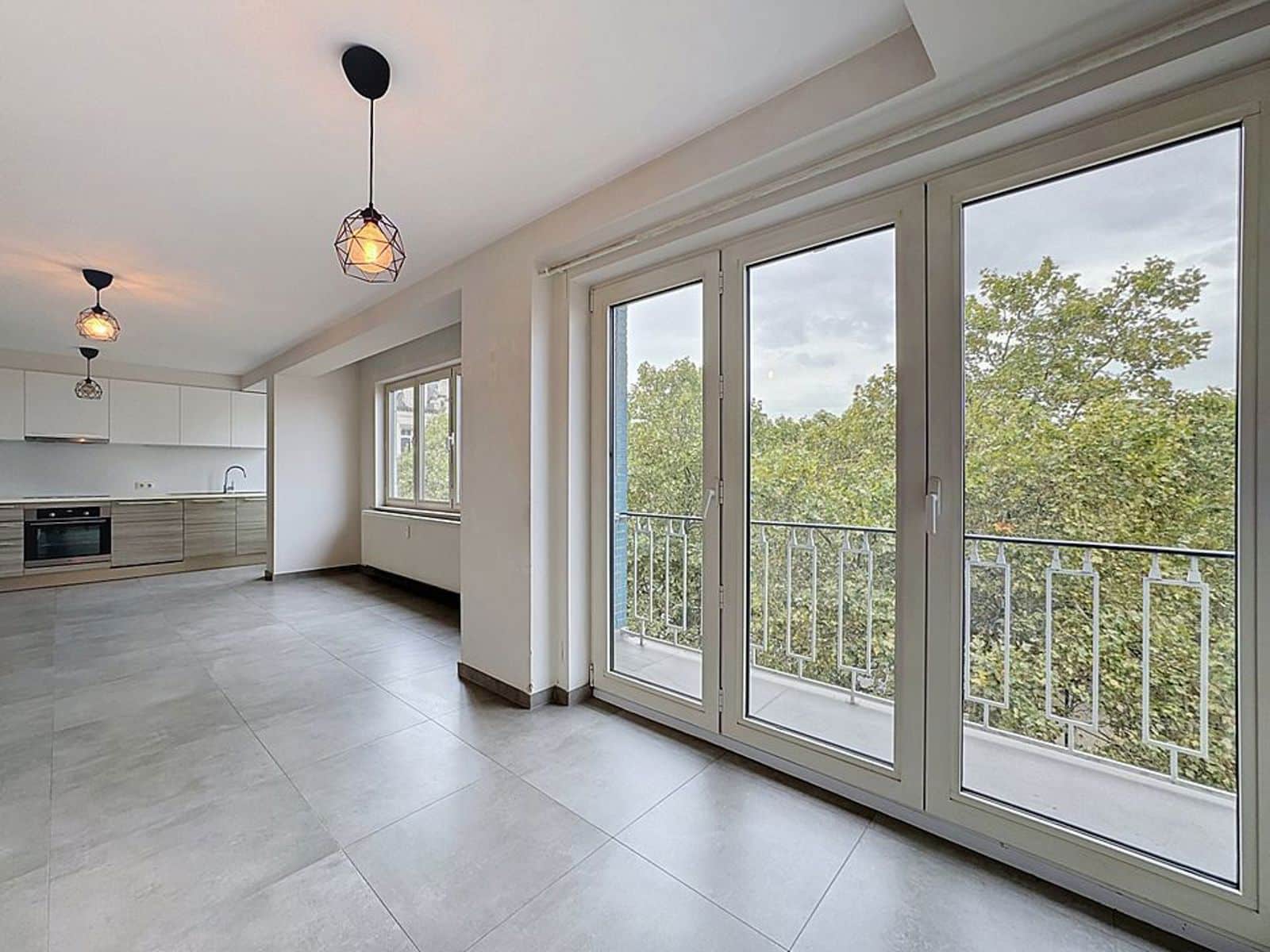 Séjour avec Cuisine Ouverte et Balcon – You Real Estate Agency Appartement moderne avec salon ouvert, cuisine équipée et balcon vue verdure.