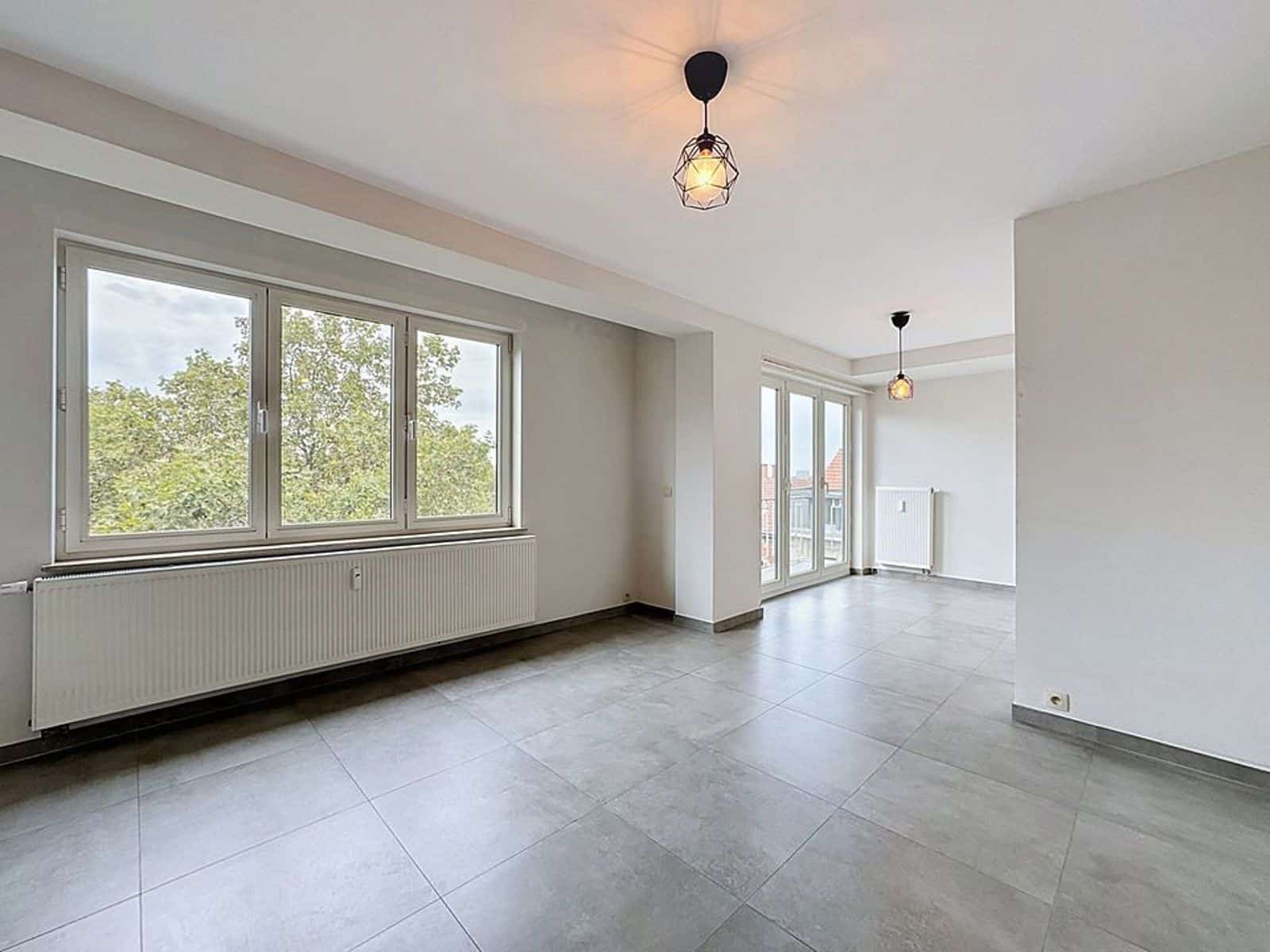 Salon Moderne et Lumineux – You Real Estate Agency Salon lumineux d’appartement moderne vide avec grandes fenêtres et radiateurs.