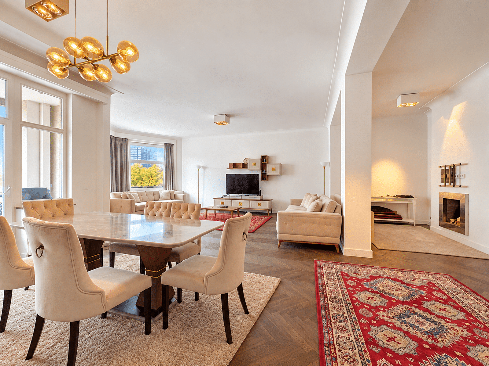 whise photo 2 Open-plan living and dining room with a marble-topped table and tufted beige chairs, a gold chandelier, and large windows letting in natural light.
