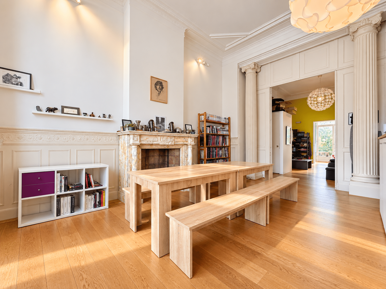 whise photo 2 Bright living room with a long wooden table and benches, marble fireplace, shelves, and tall classical columns.