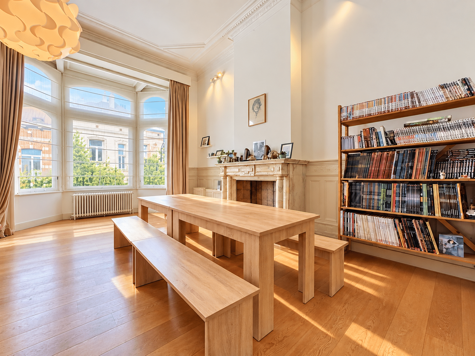 Salon Bibliothèque Lumineux – You Real Estate Agency Bright, sunlit library reading room with a large bay window, a long wooden table and benches, and a wall of magazine-filled shelves to the right.