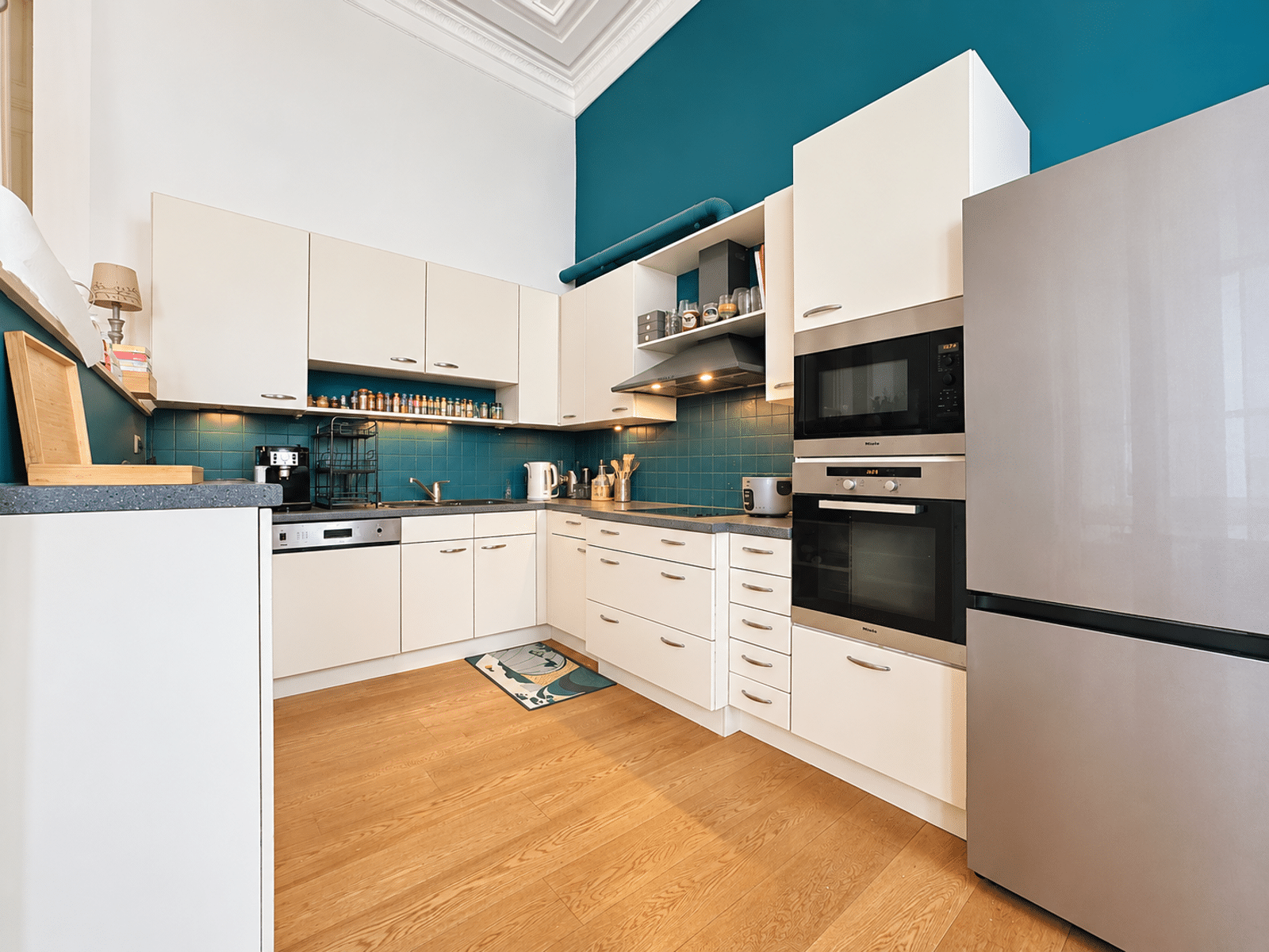 whise photo 3 Modern U-shaped kitchen with white cabinets, teal tile backsplash, and stainless steel refrigerator and double ovens on the right.
