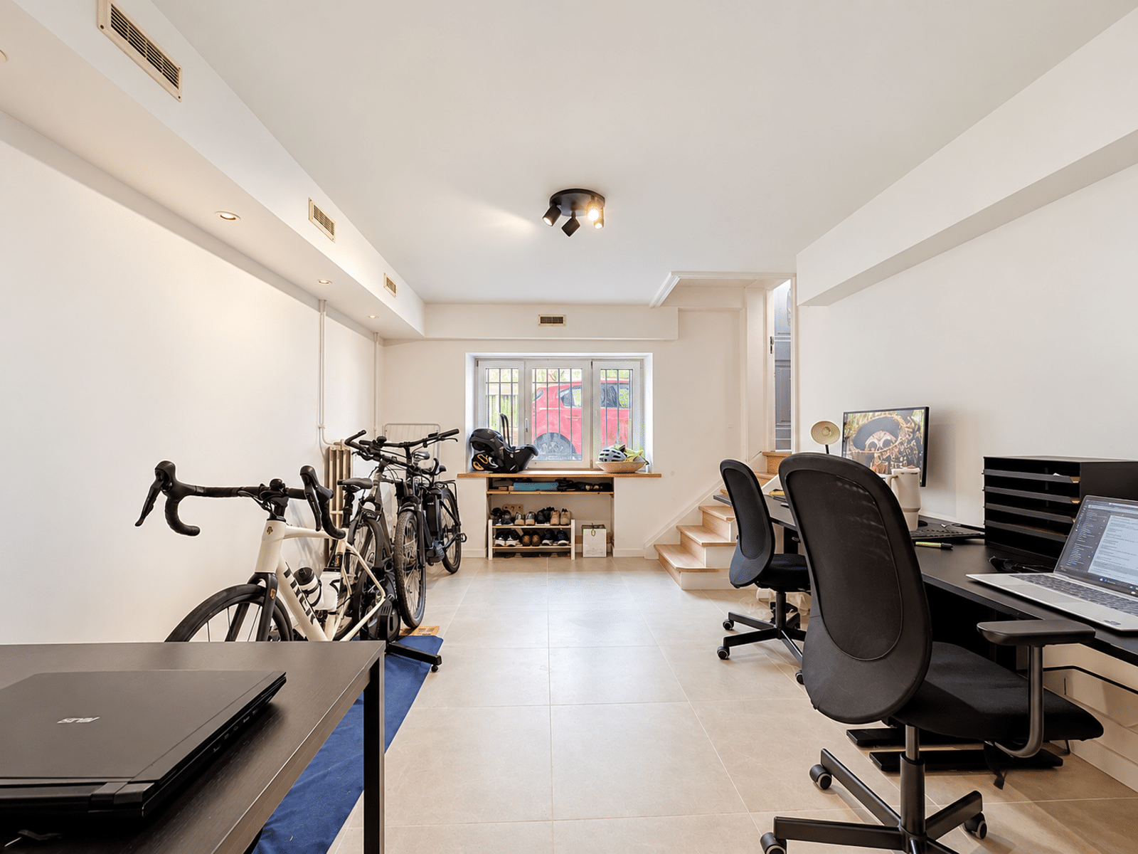 Multifunctionele ruimte met kantoor – You Real Estate Agency Een lichte werkruimte in de kelder met rechts een bureau en een stoel, fietsen die langs de linkerwand staan opgesteld, en een raam helemaal aan het einde.