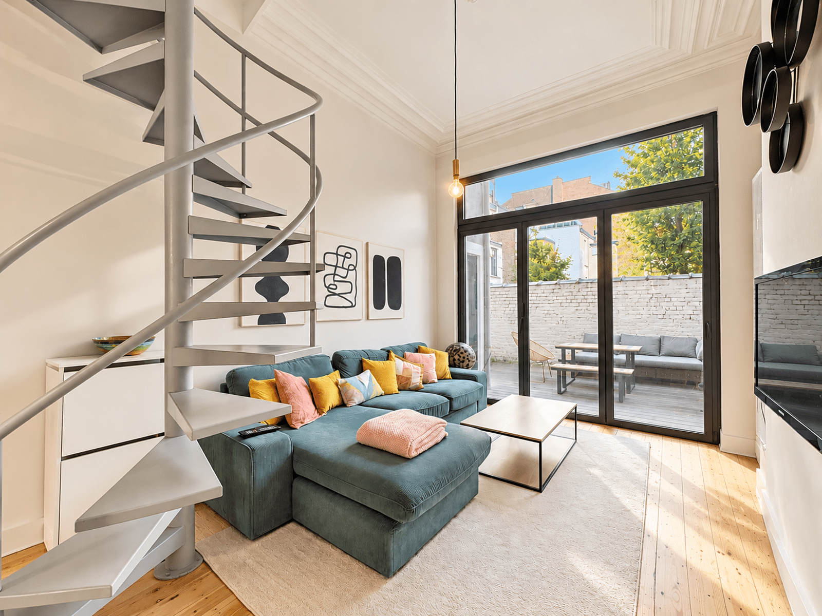 Lichte woonkamer met trap - You Real Estate Agency Moderne woonkamer met een wenteltrap, een blauwgroene hoekbank en kleurrijke kussens bij grote glazen deuren die uitkomen op een terras.