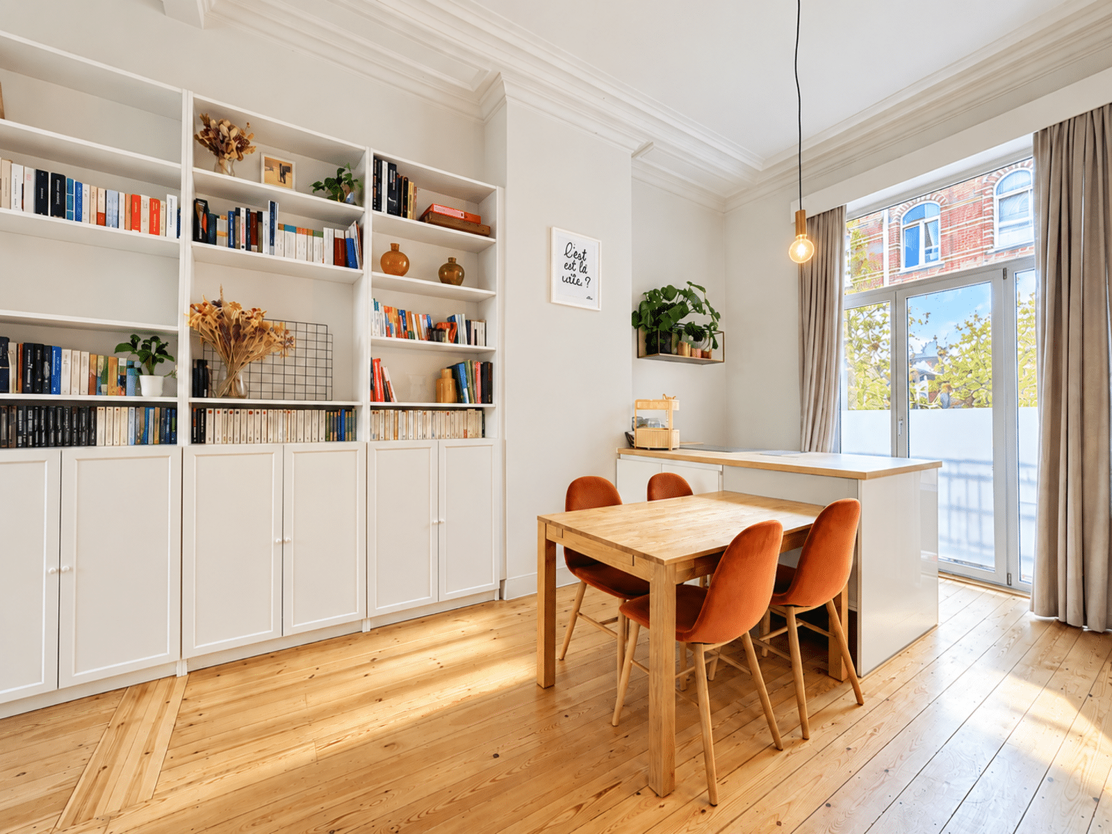 foto 3 Een lichte thuisbibliotheek met witte boekenplanken vol boeken, een houten tafel en oranje stoelen bij een groot raam.