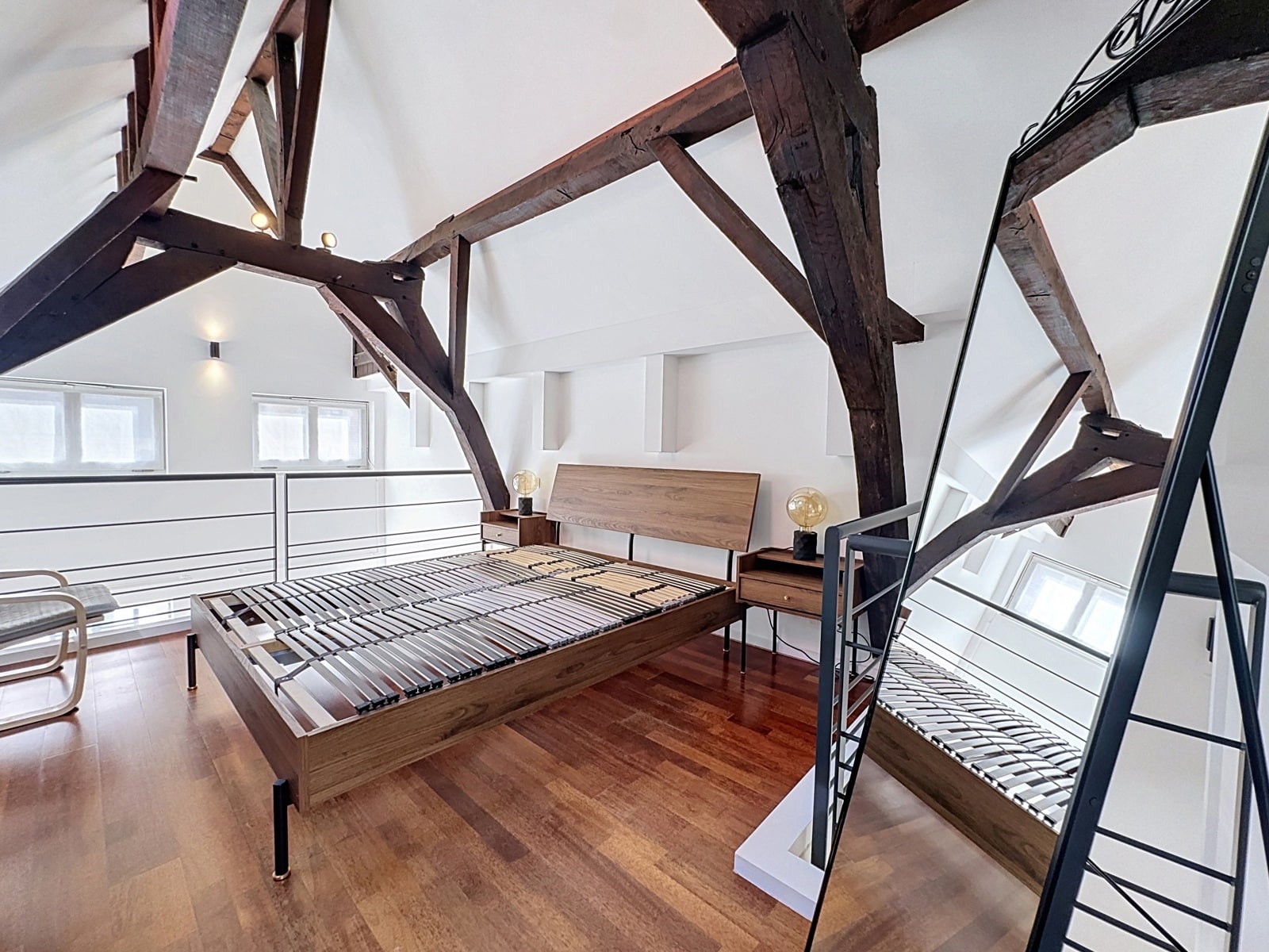 whise photo 5 Loft bedroom with dark wooden beams, a low wooden bed frame with metal slats, and matching nightstands with globe lamps on white walls and a large leaning mirror nearby.