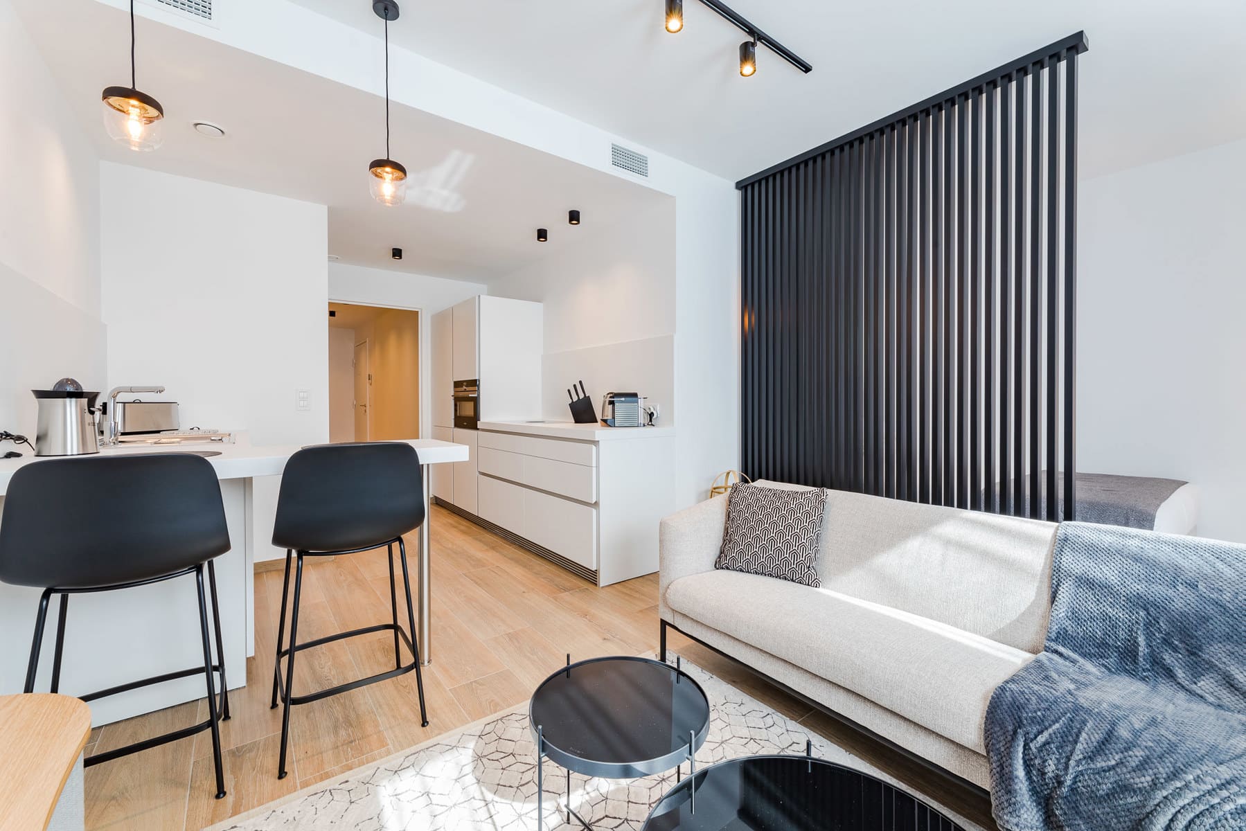 whise photo 3 Open-plan kitchen and living area with a white island, black bar stools, and a beige sofa separated by a tall black slatted divider.