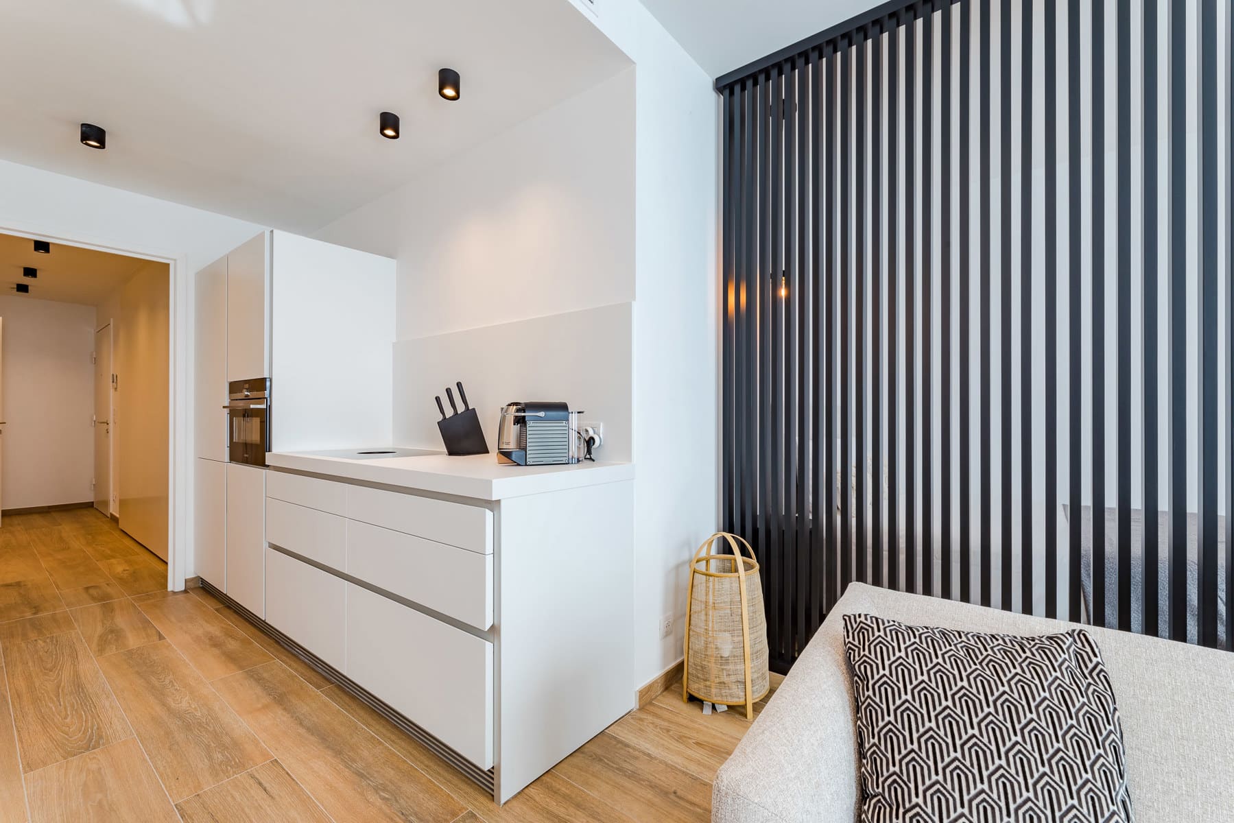 whise photo 6 Minimalist white kitchen and living area with built-in oven, knife block, and espresso machine on a white counter; black vertical slat partition on the right.