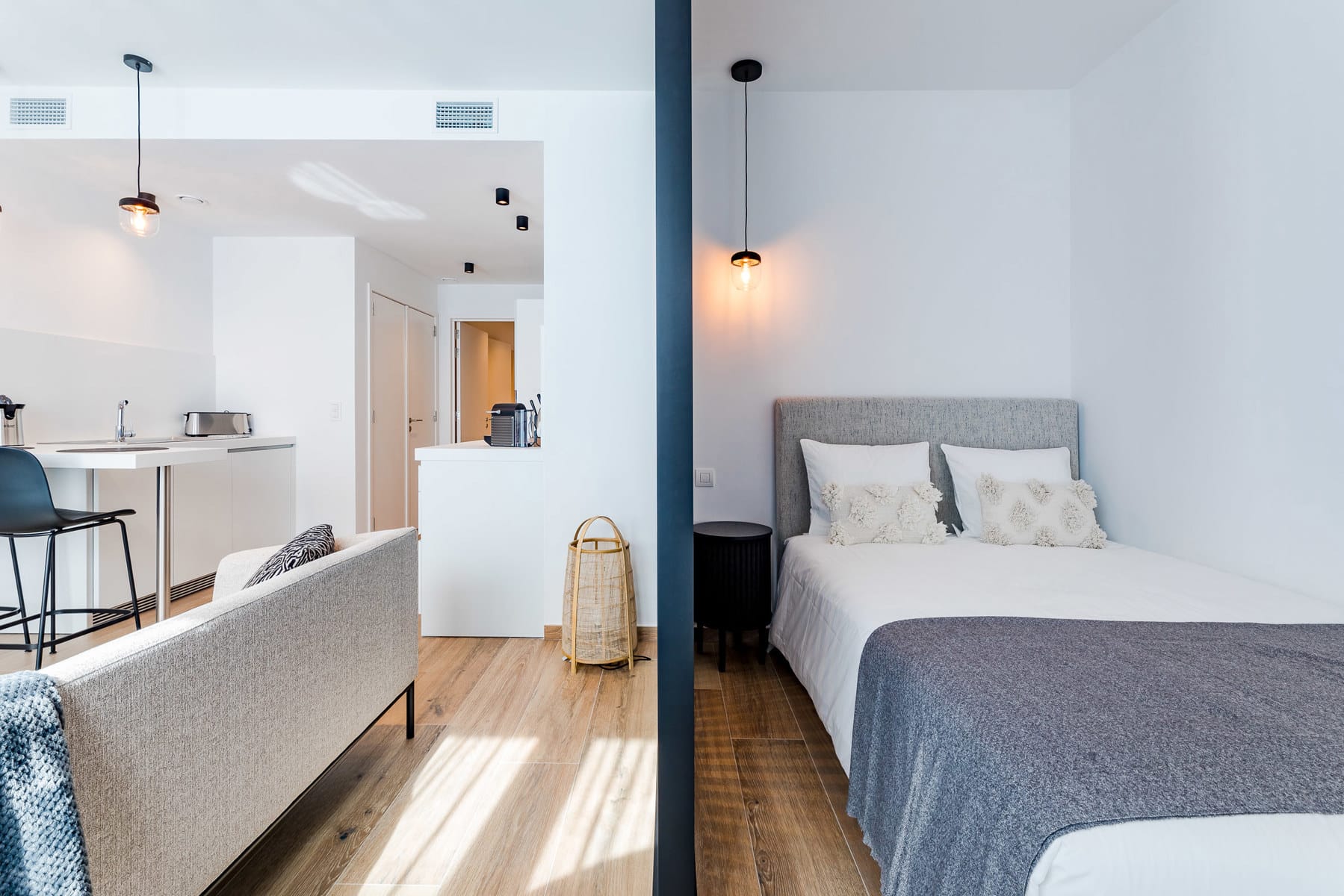 whise photo 7 Open-plan studio with a kitchen and living area on the left and a double bed on the right, split by a dark blue partition and warm pendant lights.