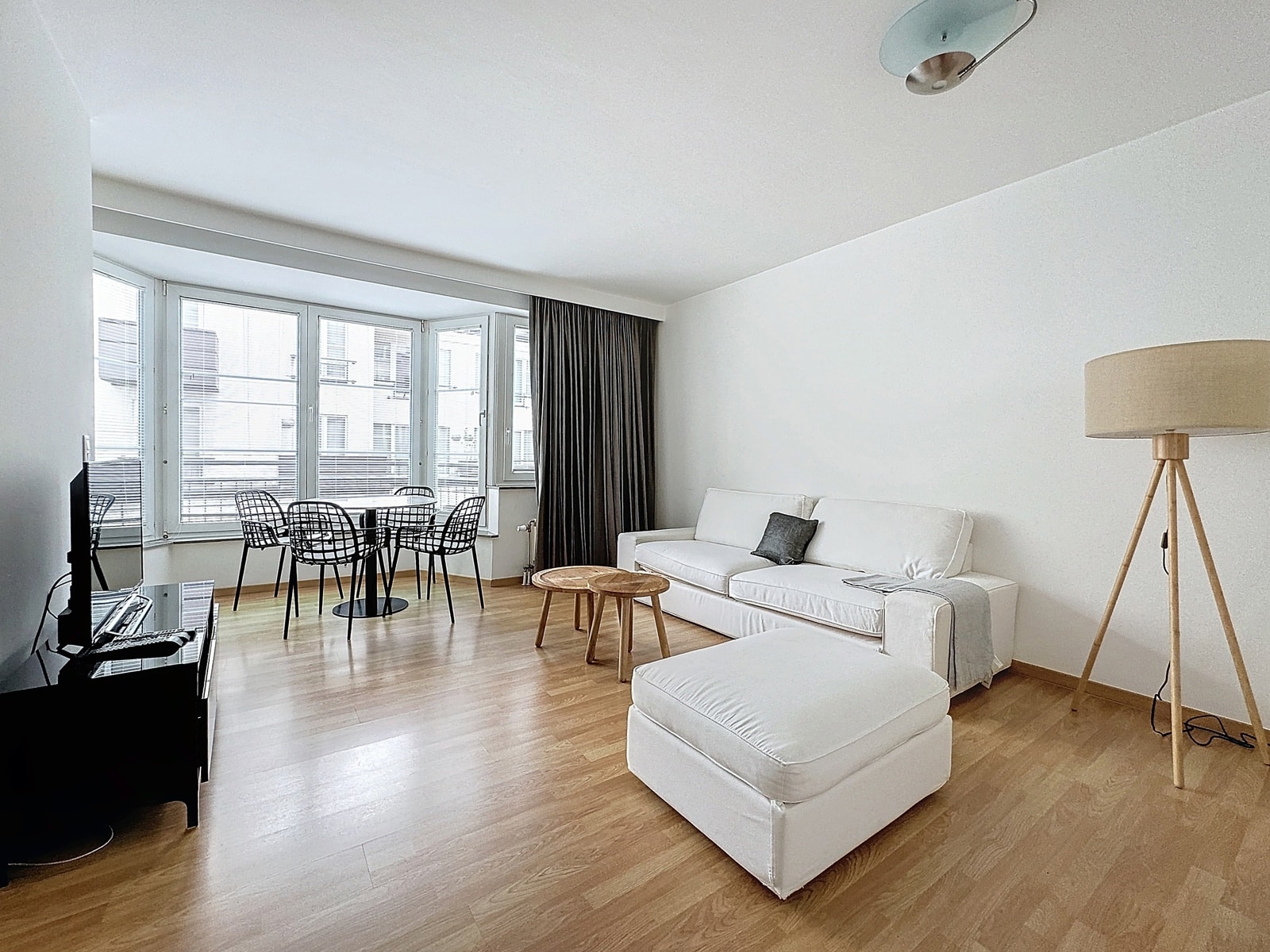 whise photo 2 A bright living room with a white sofa and ottoman, round wooden coffee tables, and a dining area next to large windows.