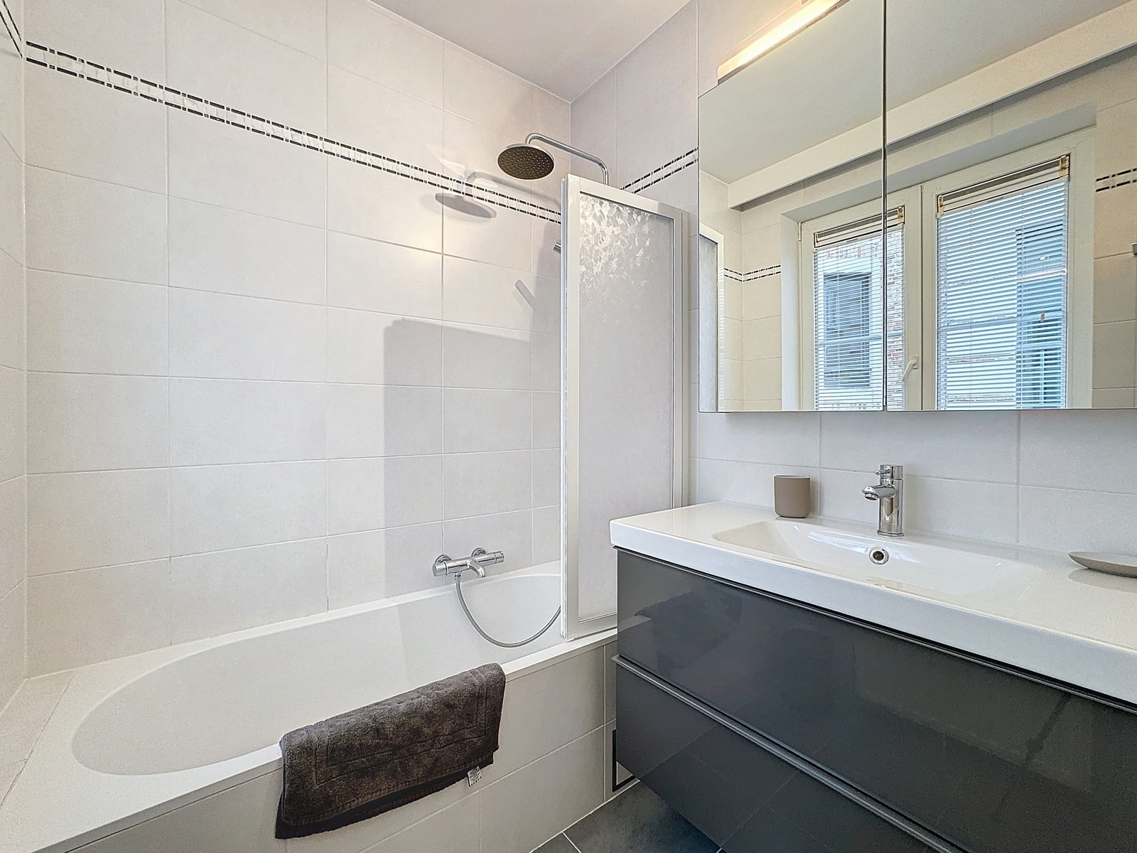 whise photo 8 Modern bathroom: white-tiled walls, a bathtub with a glass screen, and a dark gray vanity with a white sink and mirror cabinet.