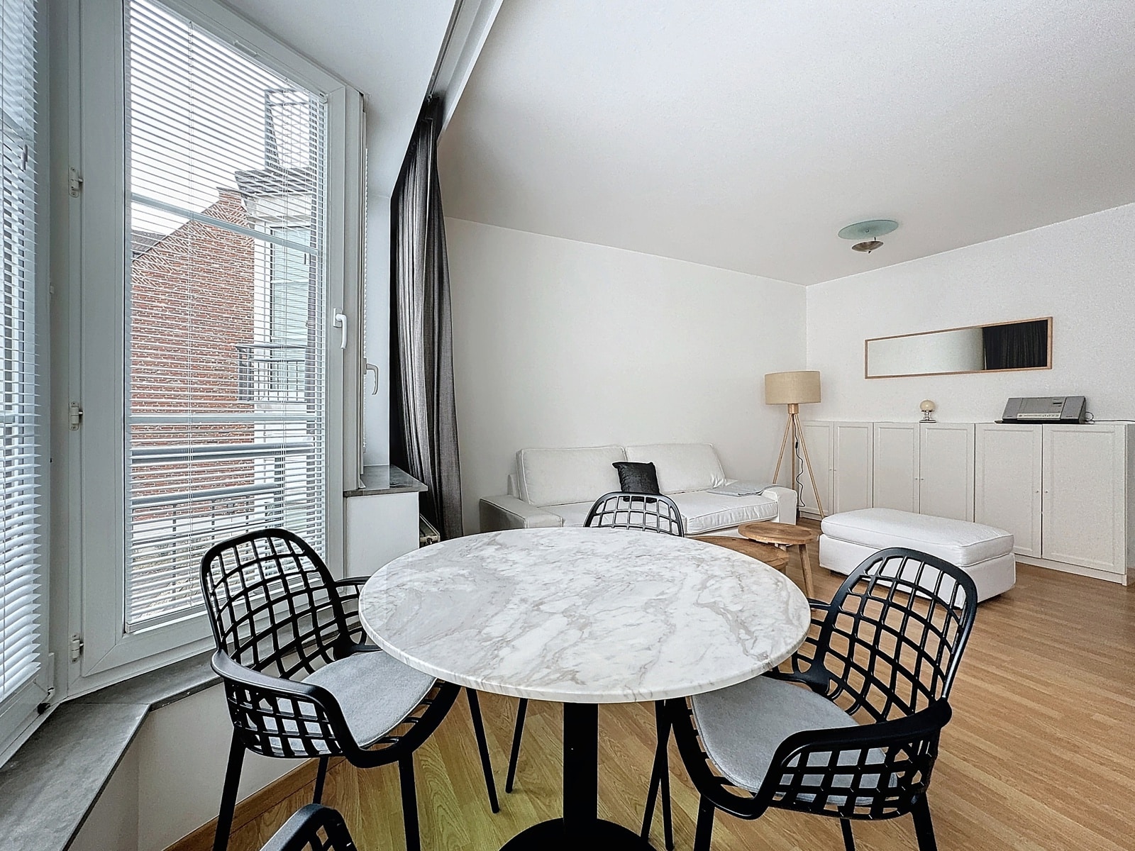 whise photo 3 A bright, open living/dining room with a round marble table and black wire chairs by a large window with blinds.