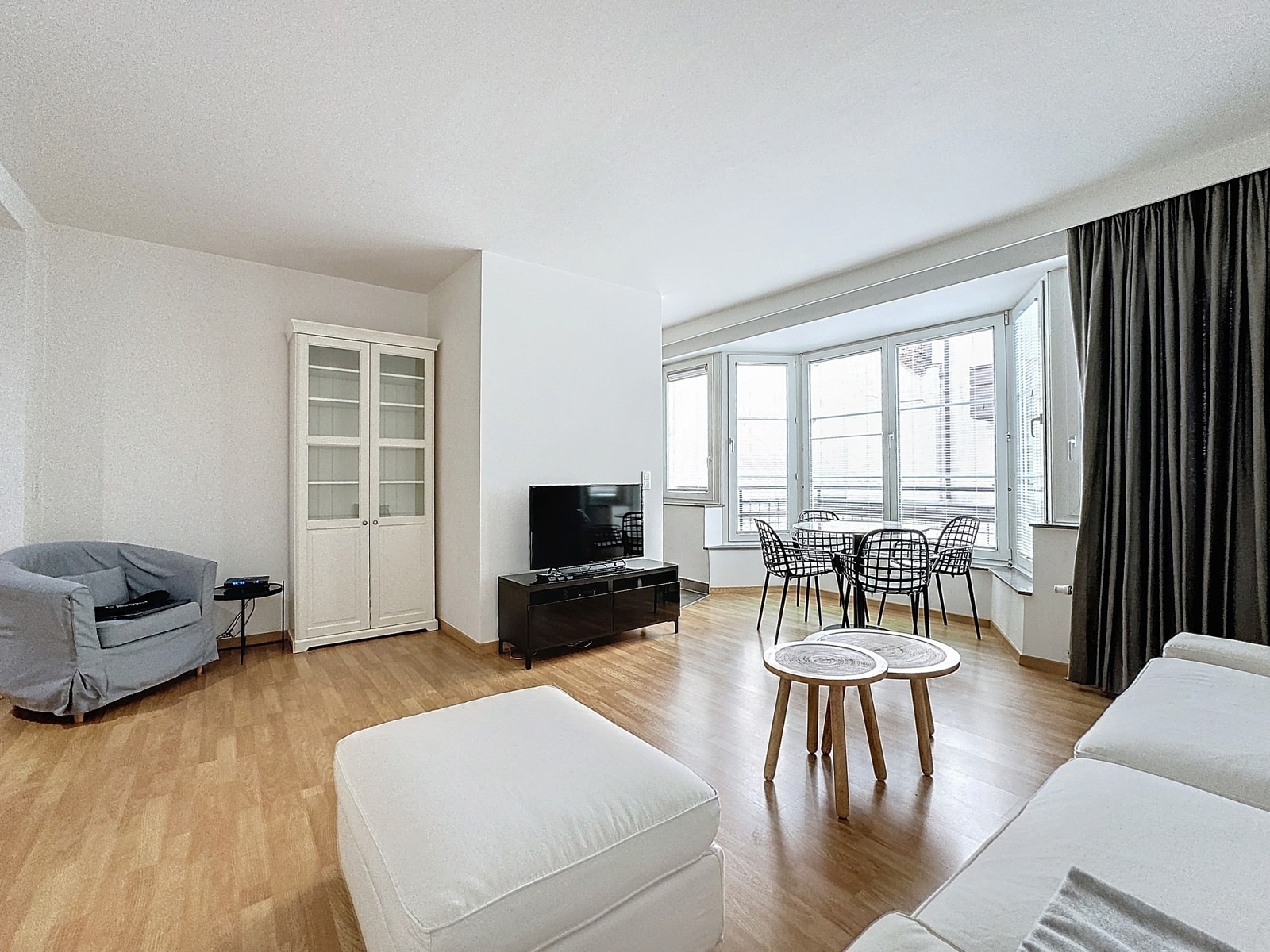 Bright Living Room with Dining Area – You Real Estate Agency A bright living room with white furniture, a TV on a black stand, a glass-front cabinet, and a bay window with a round dining table and wire chairs.