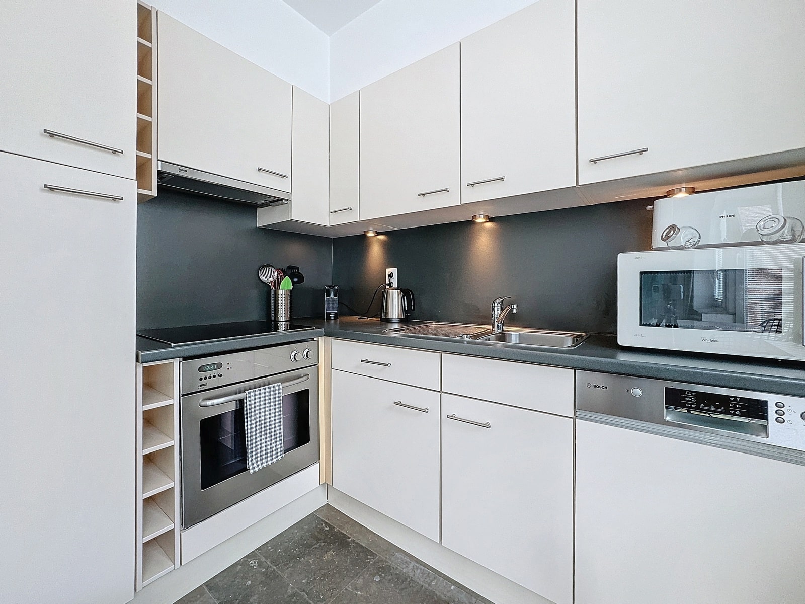 whise photo 4 Modern L-shaped kitchen with white cabinets, dark countertops, and a stainless steel oven, stove, dishwasher, and microwave, featuring under-cabinet lighting.