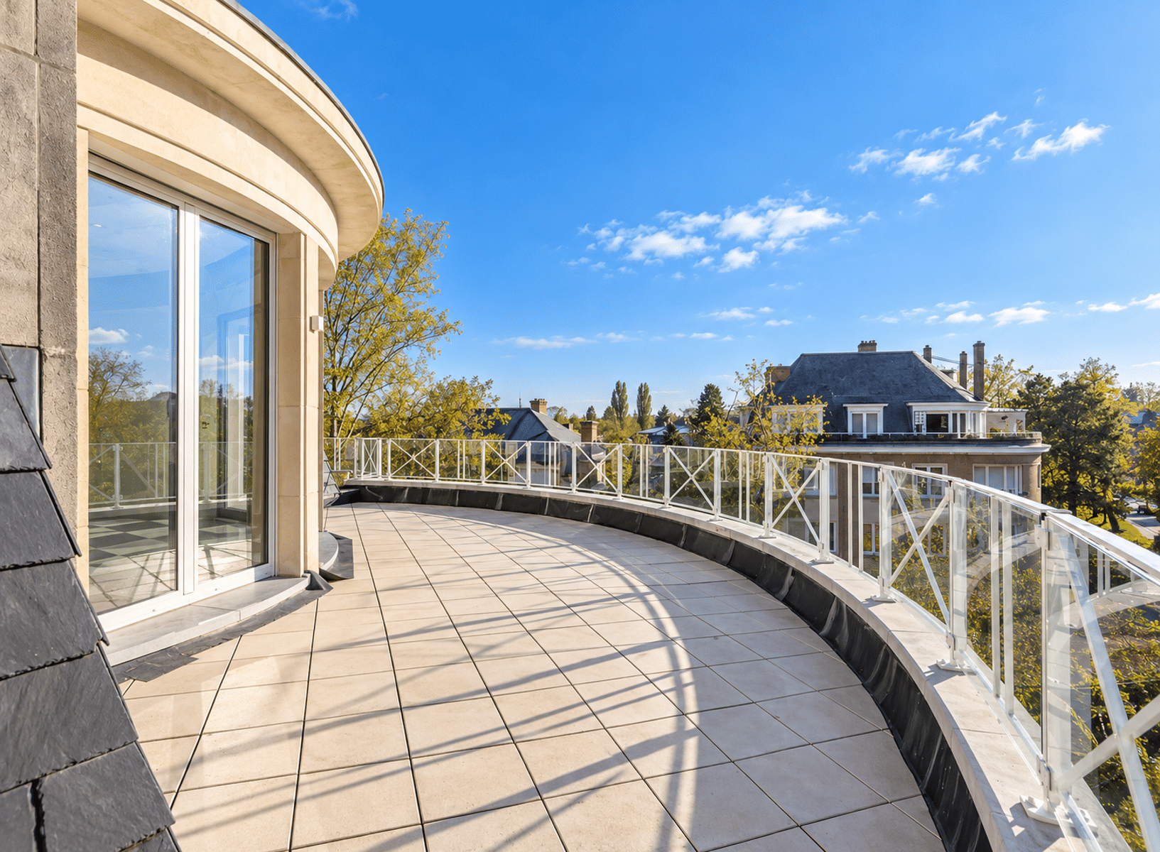 Terrasse Panoramique avec Vue Arborée – You Real Estate Agency Terrasse panoramique d’appartement moderne avec garde-corps vitré et vue résidentielle arborée.