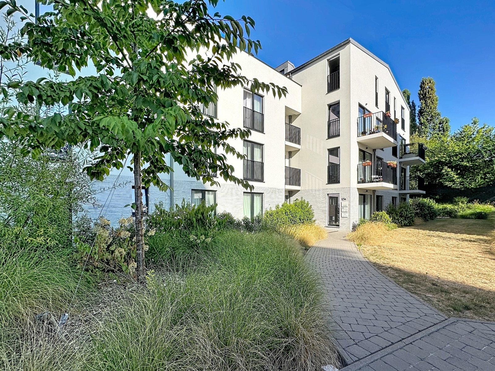 whise photo 14 A modern white apartment building with multiple balconies and a paved walkway, surrounded by greenery under a clear blue sky.