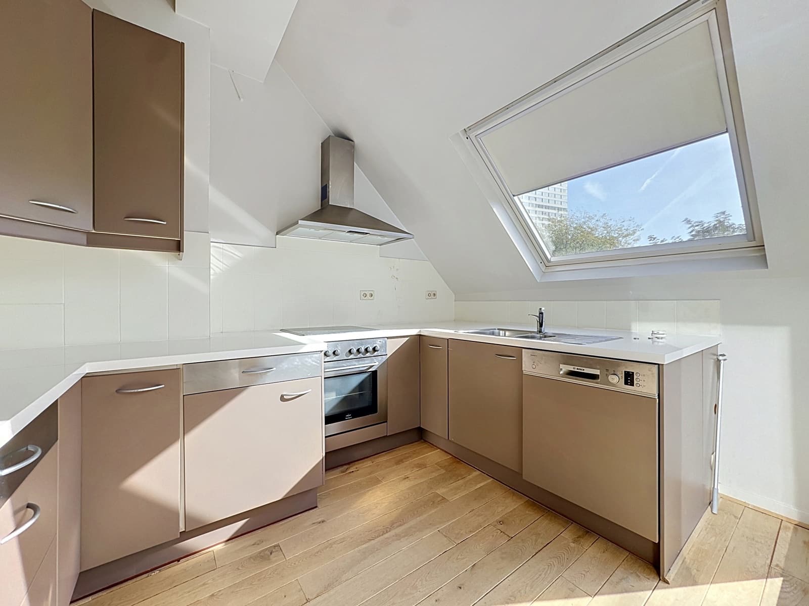 Cuisine Moderne Sous Combles – You Real Estate Agency L-shaped kitchen with brown cabinets, stainless steel oven and range hood, and a large skylight above the sink.