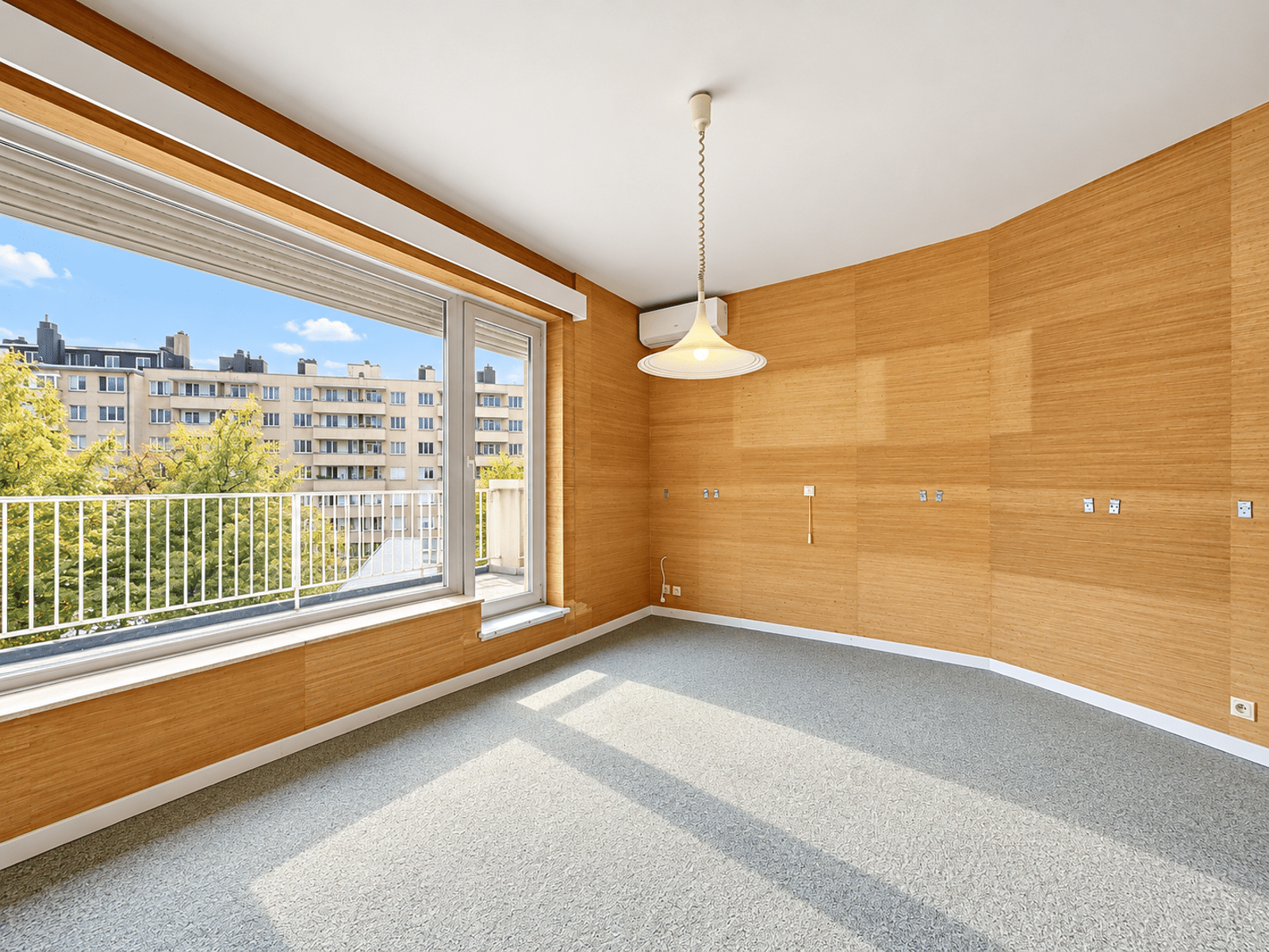 Salon Lumineux avec Balcon – You Real Estate Agency Salon lumineux d’appartement avec balcon, murs boisés et vue urbaine.