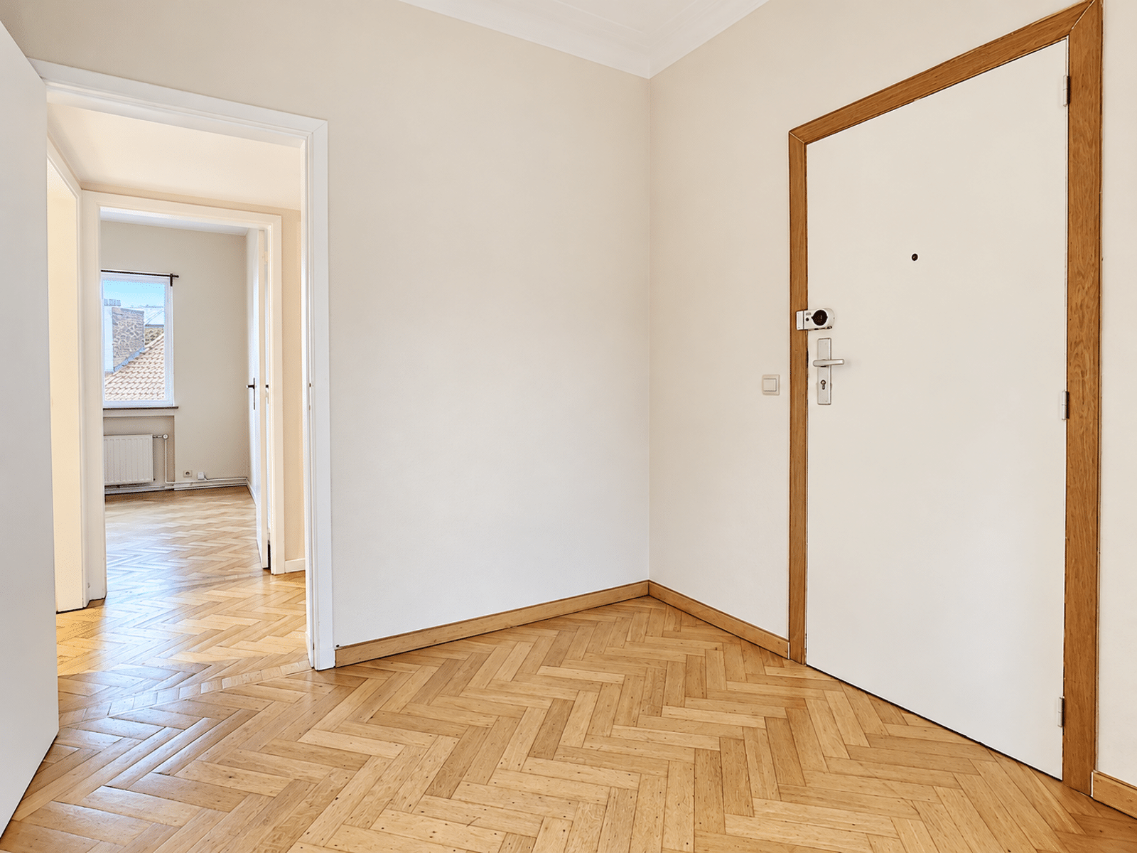 Entrée Lumineuse avec Parquet – You Real Estate Agency Entrée d’appartement lumineuse avec parquet en chevrons et porte blanche.
