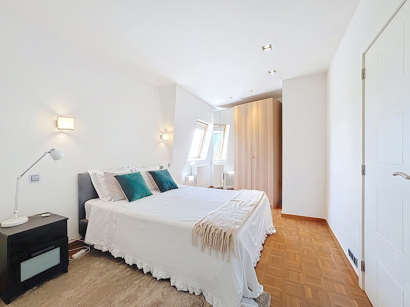 Chambre Lumineuse Sous Combles – You Real Estate Agency Chambre lumineuse d’appartement avec lit blanc, parquet, placards et fenêtres de toit.