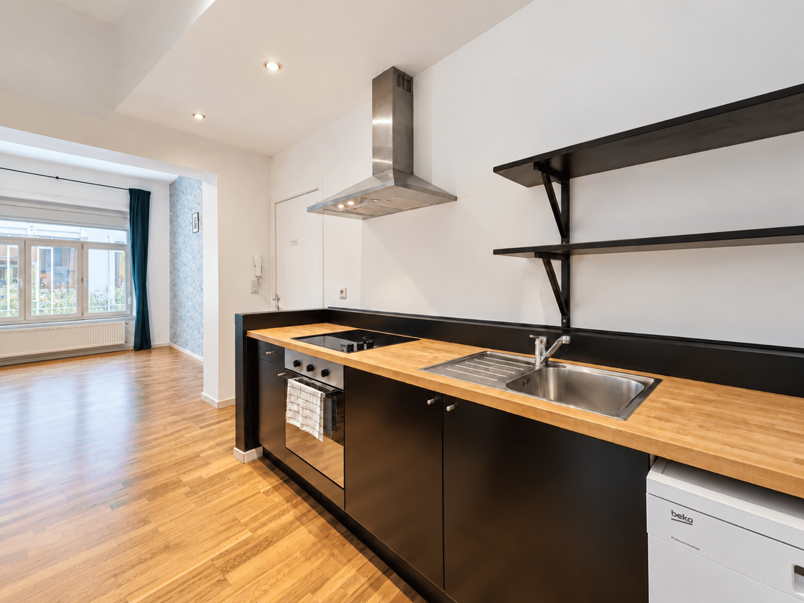whise photo 5 Modern kitchen with black cabinets, wooden countertop, stovetop and stainless steel sink along a white wall, open to a bright living area with a window and blue curtain