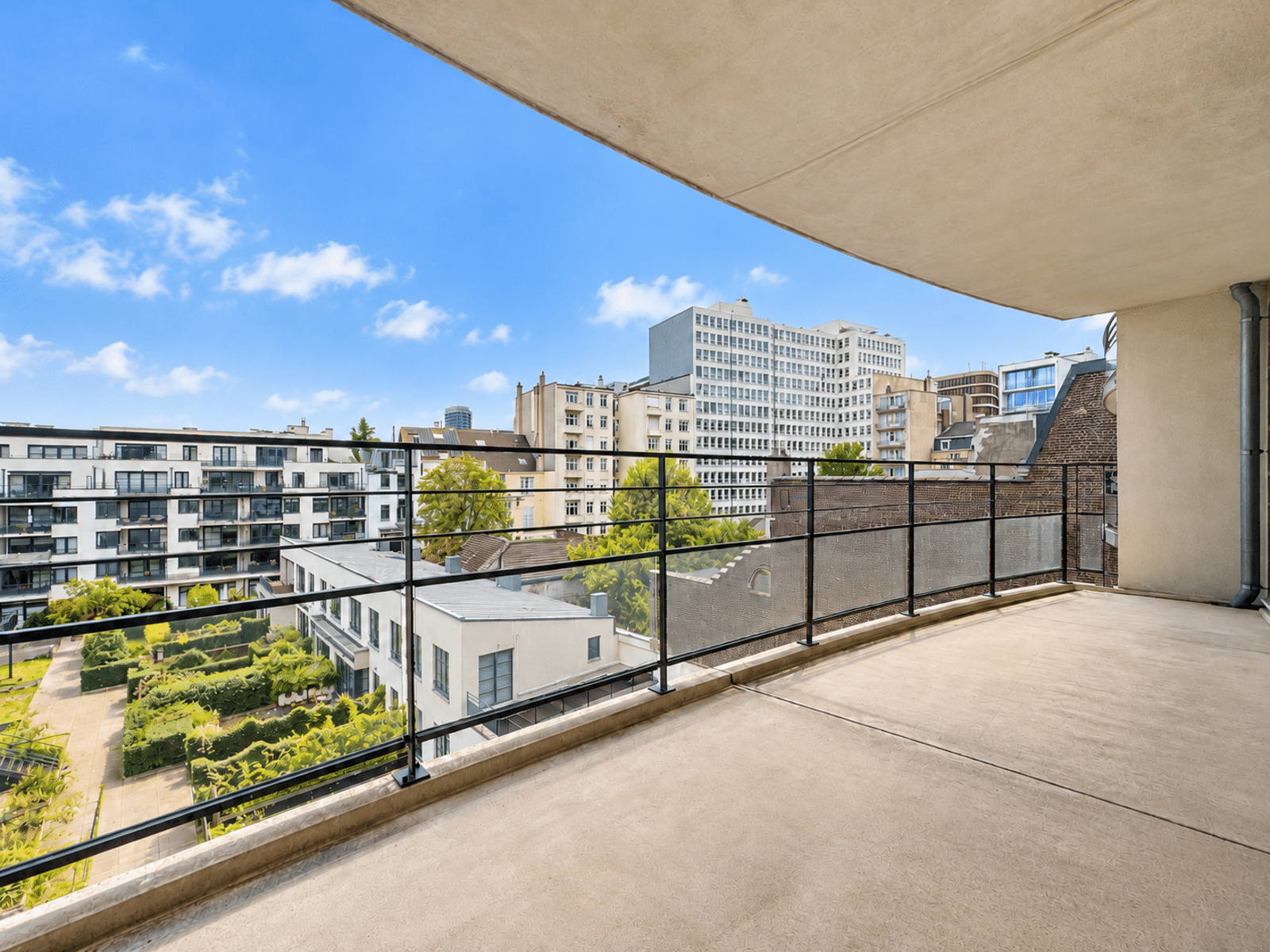 Terrasse Moderne avec Vue Urbaine – You Real Estate Agency Balcony view overlooking modern apartment buildings with a blue sky and greenery below.